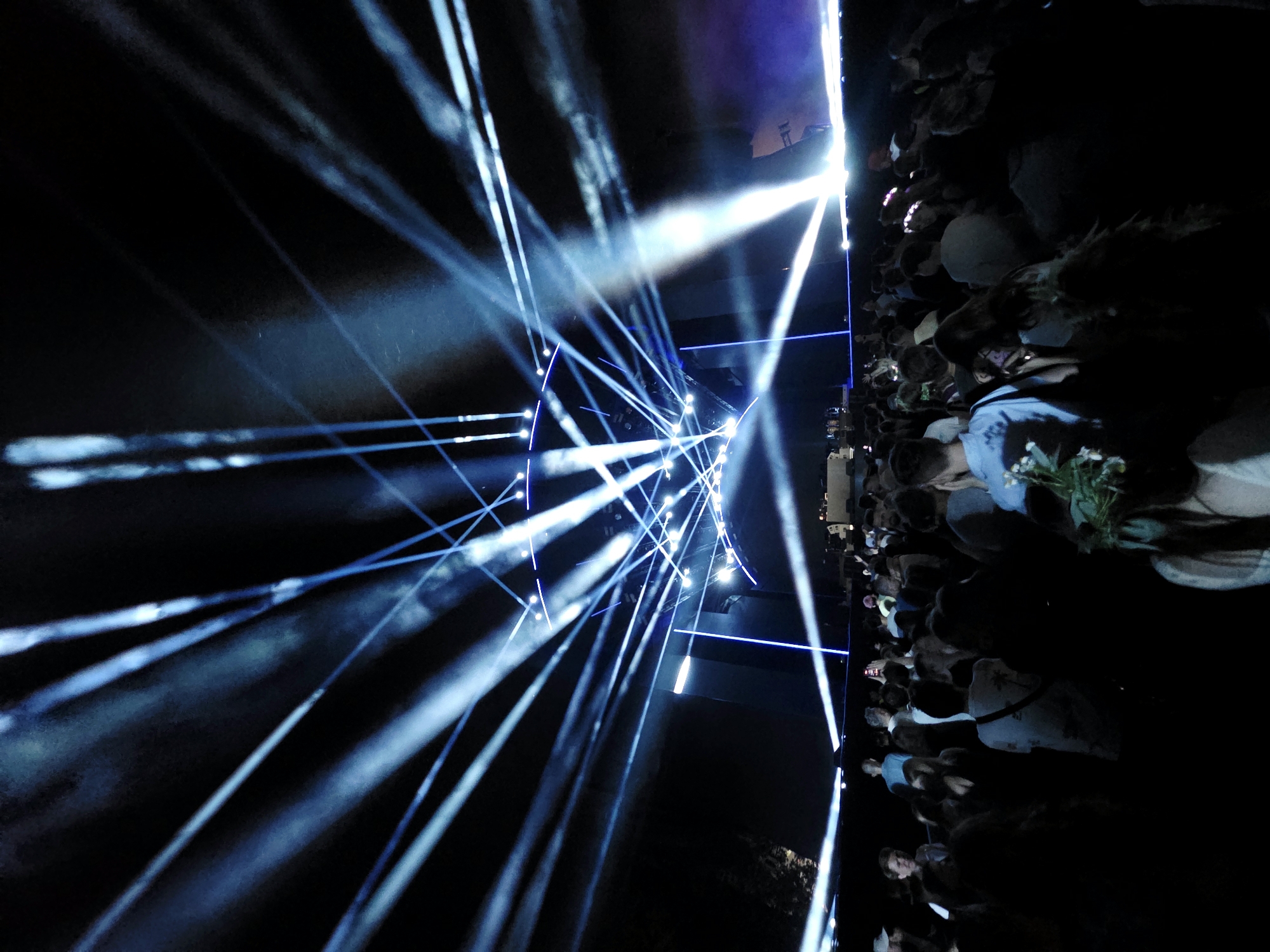 This is a nighttime photograph taken outdoors, likely at a large event, possibly a concert or festival. The dominant feature is a dramatic light show with numerous bright blue and white beams of light emanating from a stage area in the background and projecting into the dark sky. The beams create a visually striking effect, cutting through a hazy atmosphere. In the foreground and midground, a crowd of people is visible, silhouetted against the bright stage lights. Many individuals are looking towards the stage, suggesting they are attendees of the event. Some people are holding up phones, possibly to record or take pictures. The overall atmosphere is one of excitement and a shared experience, amplified by the intense lighting. The location is the city of Warsaw, Poland, at a large outdoor venue.