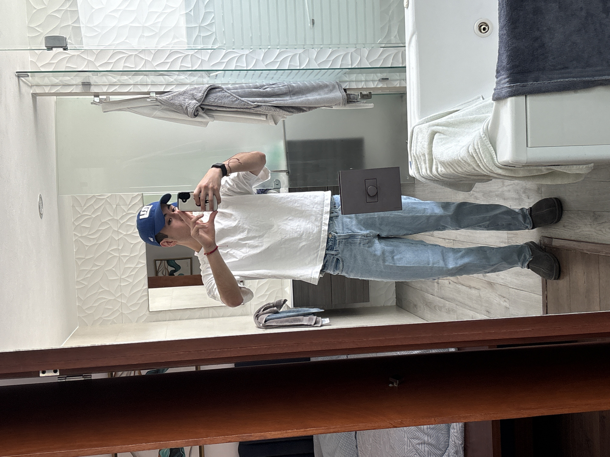 A young man stands in a bathroom, taking a selfie in the mirror. He is wearing a white t-shirt, blue jeans, and a blue baseball cap. He holds his phone in his right hand, extending two fingers toward the camera. His left hand is raised, displaying a similar gesture. A digital watch is visible on his left wrist.
The bathroom features a vanity with a dark wood cabinet, a white countertop, and a modern rectangular sink. The floor is light-colored wood-look tile. The walls are tiled with a white, textured pattern. A gray towel hangs on a glass shelf above the vanity. A toilet is visible to the right of the frame, with white towels draped over it. A stack of folded towels sits on the vanity countertop.
Based on the visual information, no specific details confirm the location is San Bernardino Tlaxcalancingo, Mexico.