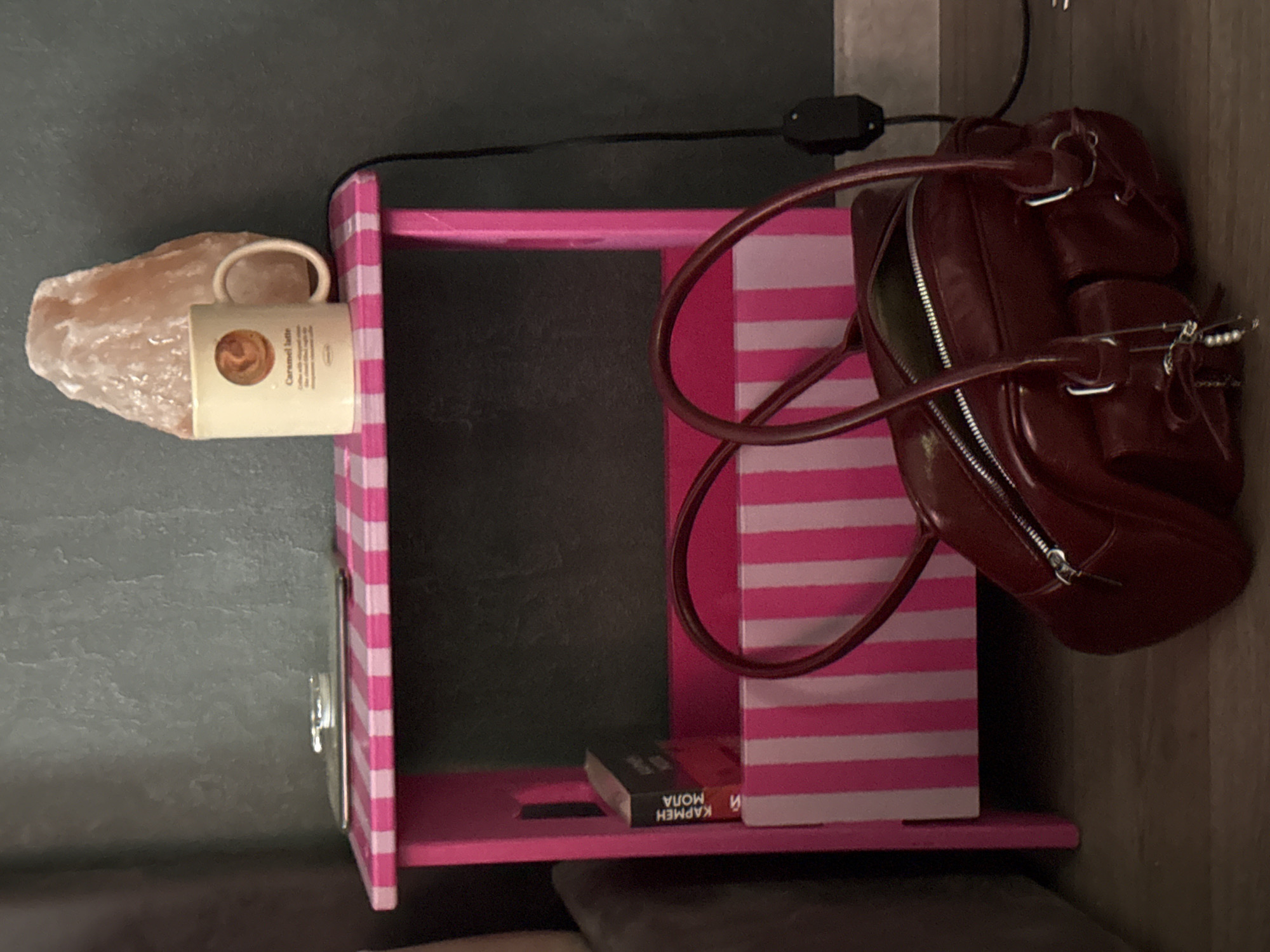 The image shows a bedside table with various items on it. On the left side of the table is a salt lamp and a mug with the words "Caramel latte" on it. On the right side of the table is a red handbag. Inside the table is a book with the title "КАРМЕН МОДА." The table itself is pink and white striped. The location is unknown.