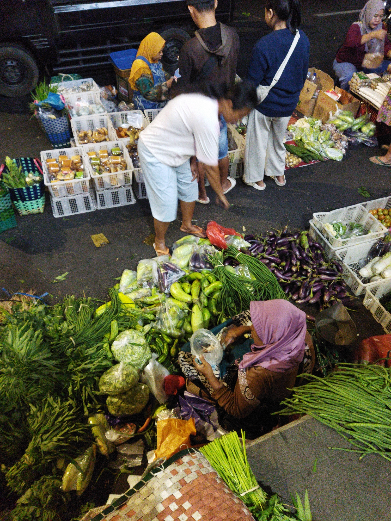 This is a medium shot, eye-level photograph taken at a street market in Semarang, Indonesia. The scene depicts a bustling outdoor market at night, with a variety of fresh produce and food items displayed for sale.
In the foreground, a woman wearing a purple headscarf and a brown long-sleeved shirt is seen from the back, interacting with a display of vegetables. She is holding a plastic bag and appears to be examining or packaging produce. To her right, a large bundle of long green beans is neatly tied with string and placed on the ground. Behind her, a variety of other vegetables, including eggplants, gourds, and leafy greens, are piled up, some in plastic bags and others loose.
Further back, to the left, several white plastic crates are filled with various food items, possibly snacks or pastries, arranged in neat rows. A woman in a yellow headscarf and a blue and green patterned apron is standing near these crates, also interacting with the market goods. She is holding what appears to be a small package.
In the background, a dark-colored truck with large tires is partially visible, suggesting the market is set up near a road. Other individuals are present in the scene, including a person in a dark hoodie and another in a blue long-sleeved shirt and light-colored pants, who are standing and observing the market activities. The ground is asphalt, typical of a street or parking area. The overall atmosphere is that of a vibrant and active night market.