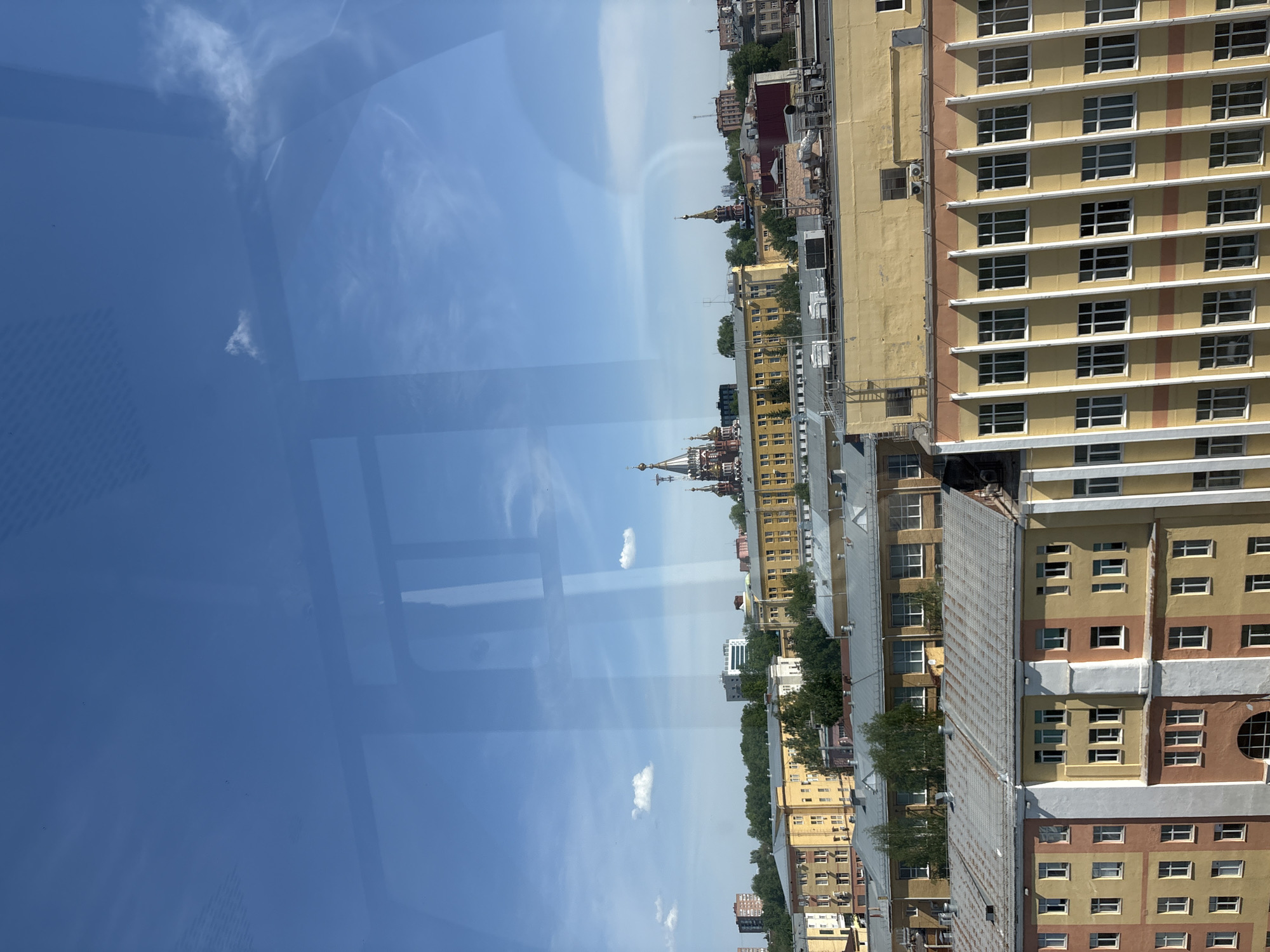 The image depicts a cityscape of Izhevsk, Russia, likely taken from an elevated position such as a high-rise building. The buildings vary in height and architectural style, with a mix of residential and commercial structures. The color palette of the buildings is predominantly yellow, beige, and brown. The sky is bright blue with a few scattered clouds. A significant portion of the image is taken up by the reflection of the sky on a window, which can be seen in the left side of the shot. In the center of the cityscape, there's a building with a distinctive Russian Orthodox church architectural style, featuring onion domes. Vegetation, primarily trees, can be seen interspersed among the buildings. There are no people visible in the shot. The overall environment appears to be urban and relatively dense.
