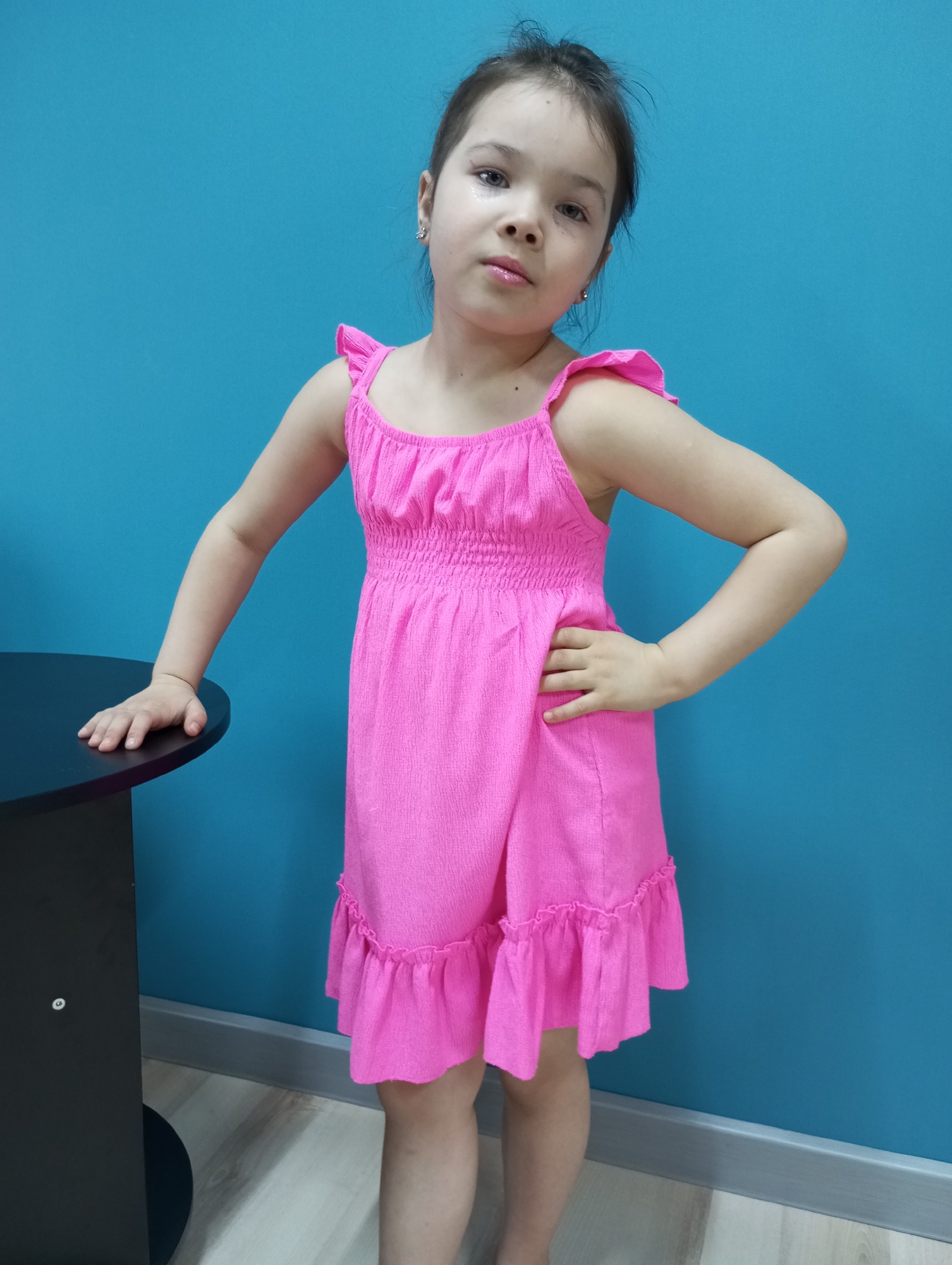 A young girl stands in front of a blue wall, posing confidently with one hand on a black table and the other on her hip. She is wearing a bright pink summer dress with ruffled straps and hem. The location is unspecified, and there is no evidence to suggest it is Pervomaysky, Russia.