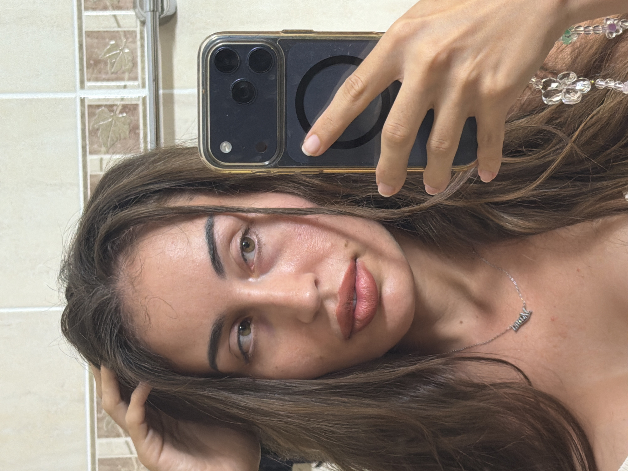 A young woman with long, brown hair is taking a selfie in a bathroom. She is holding her phone in her right hand, which is positioned in front of her face. Her left hand is resting on her head, near her hairline. She is looking slightly to her left, away from the camera. She is wearing a silver necklace with a pendant that appears to be a name. The bathroom wall behind her is tiled with a pattern of brown squares, each with a beige leaf design. A chrome towel rack is visible on the wall to the right. The scene appears to be indoors, likely in a residential setting.