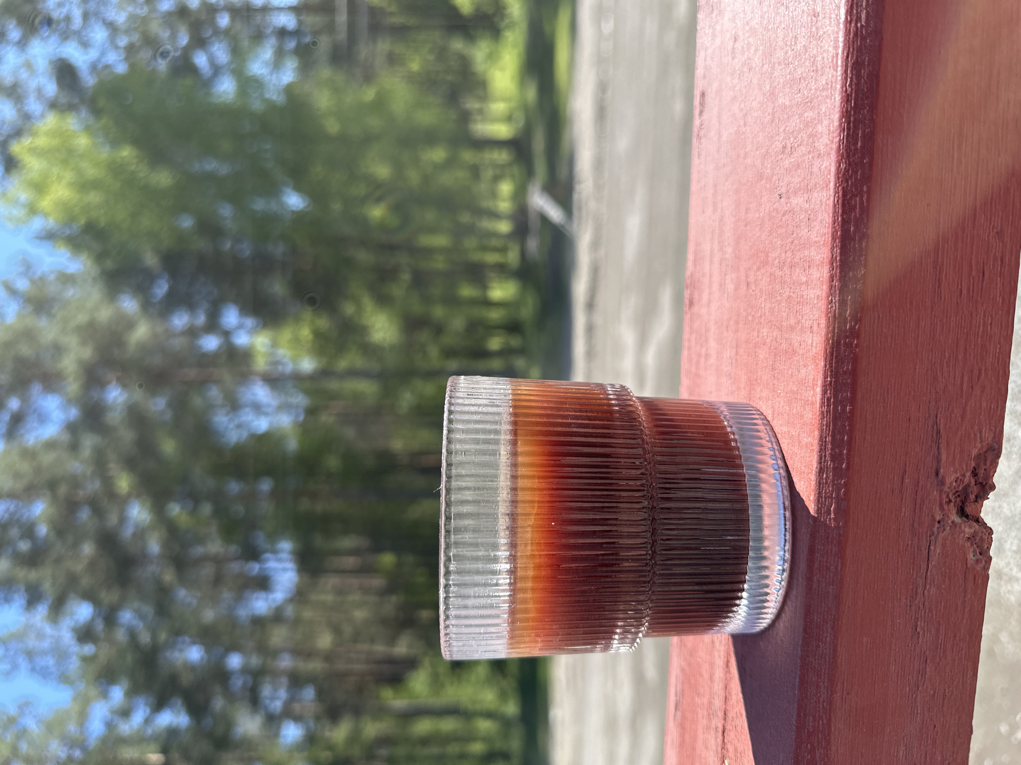 The image shows a glass of dark liquid on a red wooden surface, possibly a ledge or windowsill. The glass has a clear, ribbed design. The liquid appears to be layered with varying shades of brown. In the background, blurred trees and sky are visible, suggesting an outdoor setting. The presence of trees indicates a natural environment or a park-like area. There is no explicit information in the image indicating the location is Askat, Russia, or about any people, actions, or interactions.