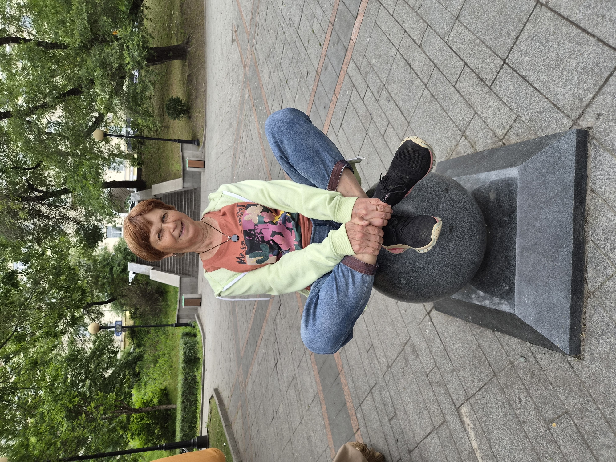 A woman with short auburn hair is seated in a lotus position on top of a large, dark gray sphere, which is mounted on a faceted dark gray base. She is wearing a light green hooded jacket over an orange t-shirt with a graphic print, blue jeans, and black athletic shoes with pink accents. She is smiling and looking at the camera. Her hands are clasped in her lap.
The scene takes place outdoors in what appears to be a public park or square with a paved ground made of gray stone tiles. In the background, there are trees with lush green foliage, stone stairs leading up a grassy slope, and lampposts. A building with windows is visible behind the trees. The overall environment suggests a pleasant, possibly temperate day. The location is identified as being in the city of Vladivostok, Russia.