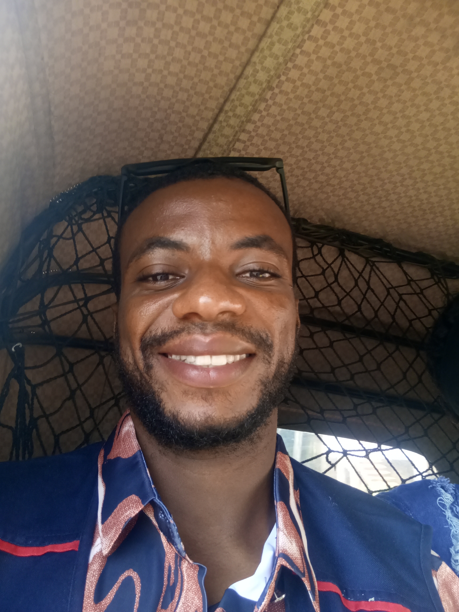 In the city of Jos, Nigeria, a man of African descent, with dark skin and a beard, smiles broadly while taking a selfie. He wears a blue shirt with a colorful patterned collar. The background shows a checkered fabric, likely the interior of a vehicle, and a black net. The lighting suggests it is daytime.