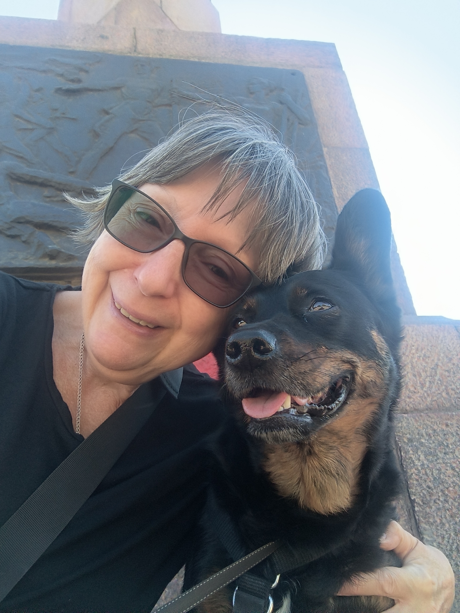 The image is a selfie of a woman and her dog. The woman is in the foreground, smiling and wearing sunglasses and a black shirt. The dog, a black and tan breed, is leaning into the woman and appears happy, with its tongue out. Behind them, a stone structure with a bas-relief carving is visible. The sky is clear and bright. The location in Mykolaiv, Ukraine, is not specified.