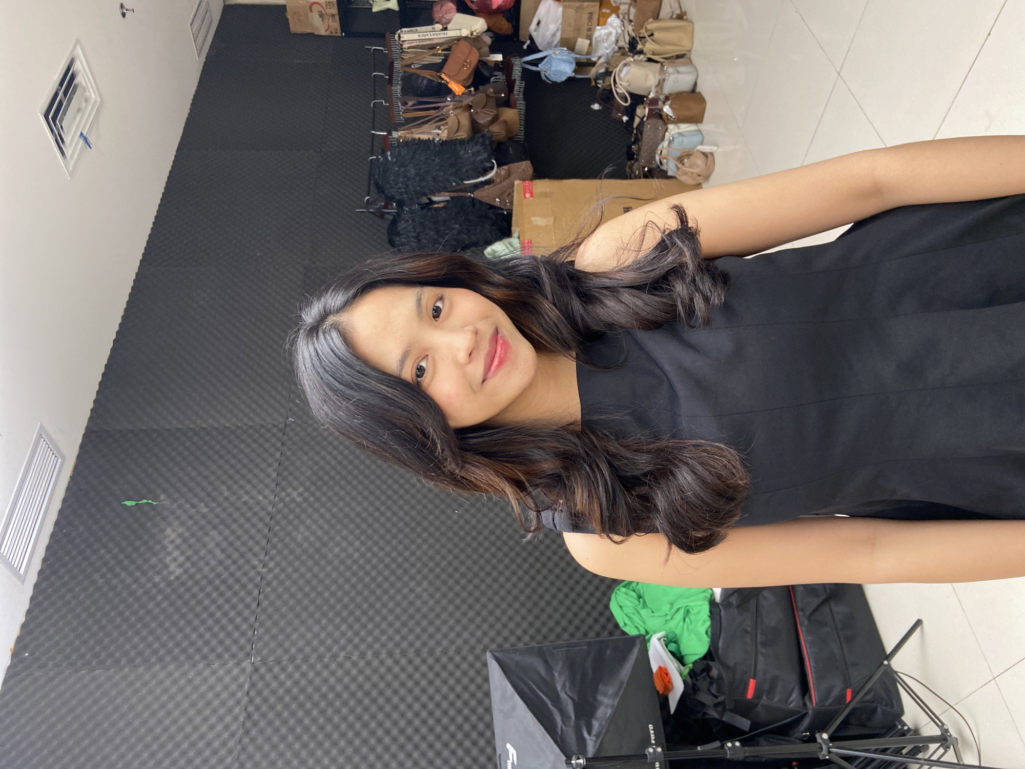 A woman with long, dark wavy hair is in the center of the frame, smiling at the camera. She is wearing a black sleeveless dress. Behind her is a wall covered in dark gray soundproofing foam. To the right and above her are numerous handbags on shelves. Below her, there are equipment bags. Based on metadata, the location is in North Jakarta, Indonesia.