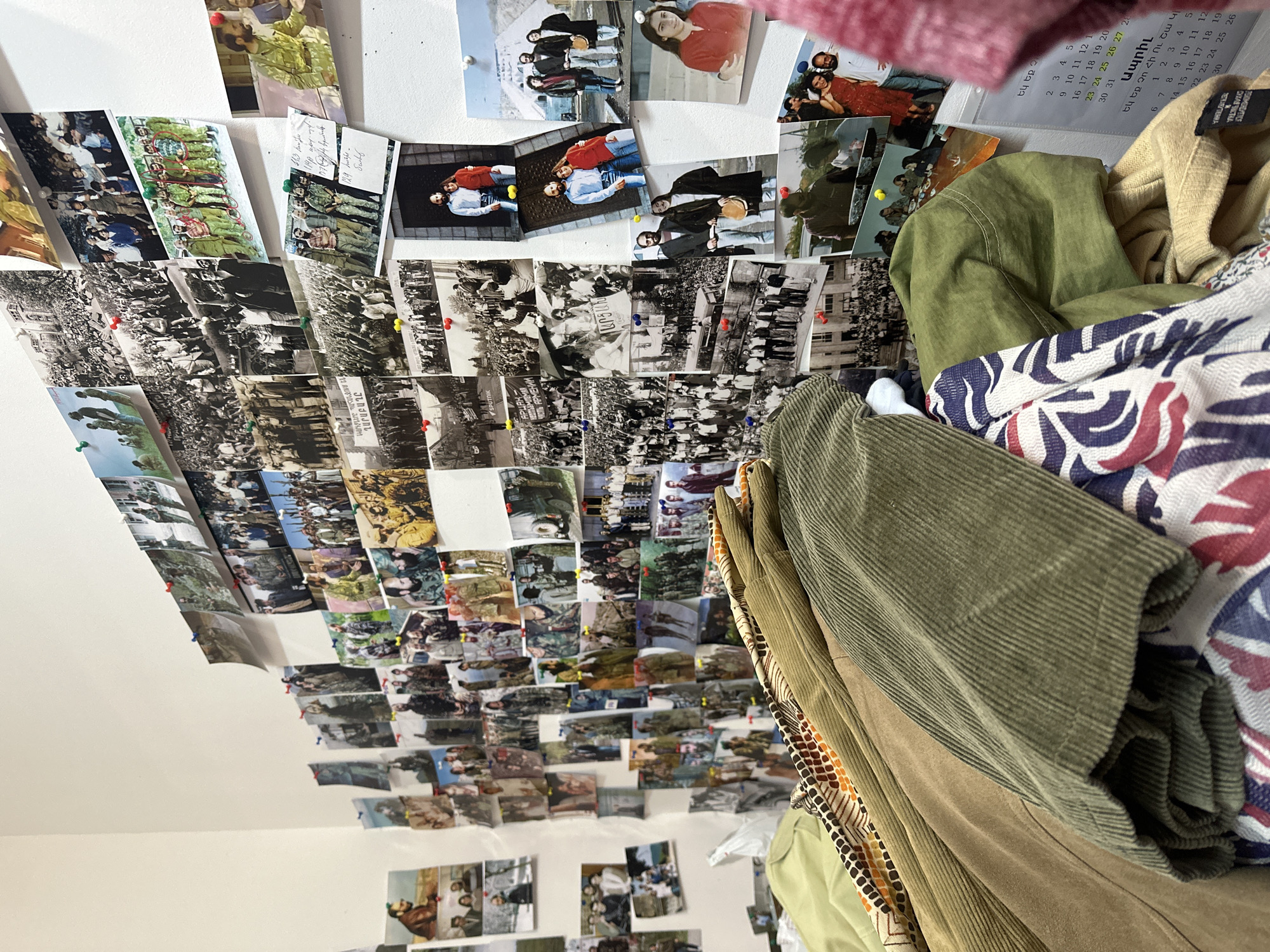 A white wall is densely covered with pinned photographs, predominantly black and white, with some in color. A calendar with Armenian text is visible in the upper right, partially obscured by clothing. A pile of assorted clothing items in various colors and textures sits against the wall. The location, Yerevan, Armenia, is inferred from the Armenian text on the calendar.