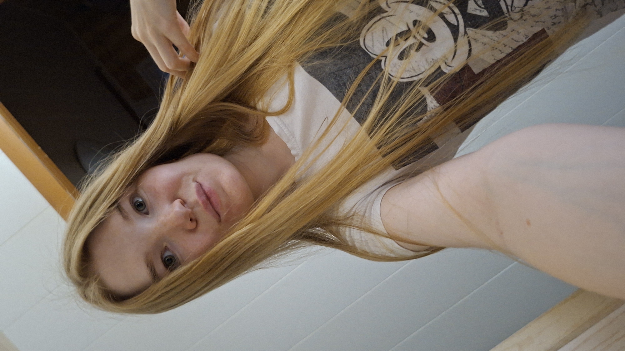 A young Caucasian woman with long, blonde hair and fair skin is taking a selfie indoors. She is wearing a white t-shirt with a graphic print that includes a stylized image of Mickey Mouse. Her left arm is visible in the foreground, and her right hand is touching her hair. The background consists of a white paneled wall and a wooden door frame. The lighting suggests an indoor setting, possibly a home. The image does not provide any information about the city of Большой Букор, Russia.