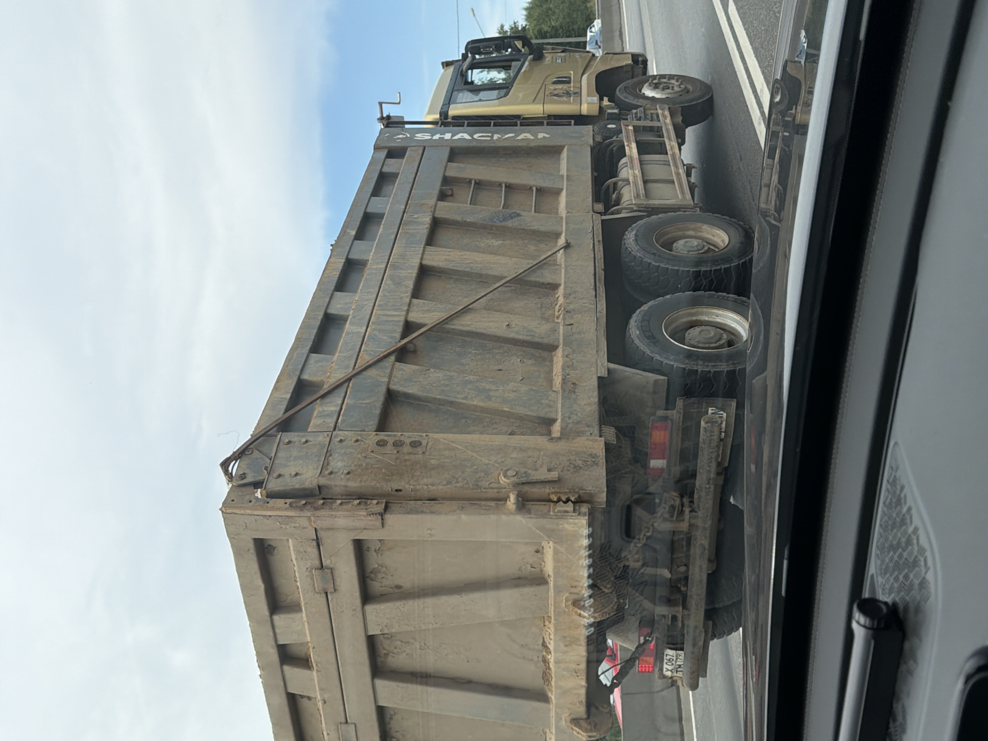 A dirty, gold-colored dump truck with a large, weathered cargo bed drives on a highway in Radumlya, Russia. The truck has the brand name "SHACMAN" written vertically on the side of the cab. Its tires are caked with dirt, and the cargo bed is full of dirt. The truck has multiple axles and large, rugged tires. In the foreground, a black dashboard with a small black device is visible, suggesting the photo was taken from inside another vehicle. The background shows a cloudy sky and a few trees along the side of the road. A red car is partially visible ahead of the dump truck. The license plate of the dump truck is visible and reads "X 067 TM 179" which is a Russian license plate.