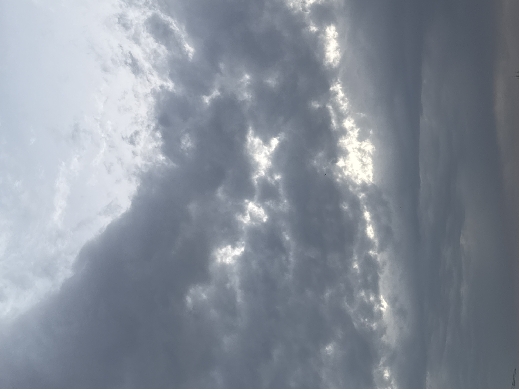 This is a photograph of a cloudy sky, likely taken from an elevated viewpoint in Bishkek City, Kyrgyzstan. The image is dominated by various shades of gray clouds, with patches of bright white where sunlight is breaking through. Some of the clouds appear thick and dark, suggesting the possibility of impending rain, while others are thinner and more diffused. In the lower left corner, a portion of a construction crane is visible, indicating an urban or developing area. The overall impression is of a dynamic sky with dramatic lighting.