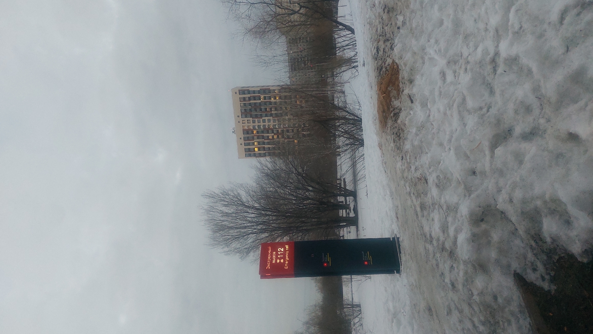 On an overcast day, a landscape depicts a snow-covered area in what is likely Novosibirsk, Russia. A tall residential building with many lit windows stands in the background. A red emergency call box labeled "Экстренный вызов 112 Emergency call" is prominently displayed. Bare trees surround the call box and building. The ground is covered in snow and dirt.