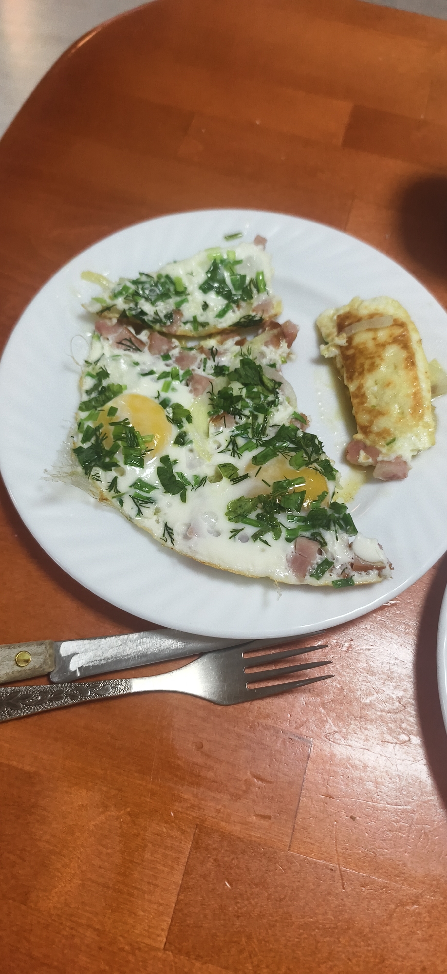 A white plate containing fried eggs with green herbs and small pieces of meat sits on a wooden table. A fork and knife rest beside the plate on the table. Part of another white plate is visible in the lower right corner of the image. The location is unknown and cannot be determined from the image. The city of Фоки, Russia cannot be identified as the location based on the provided image.