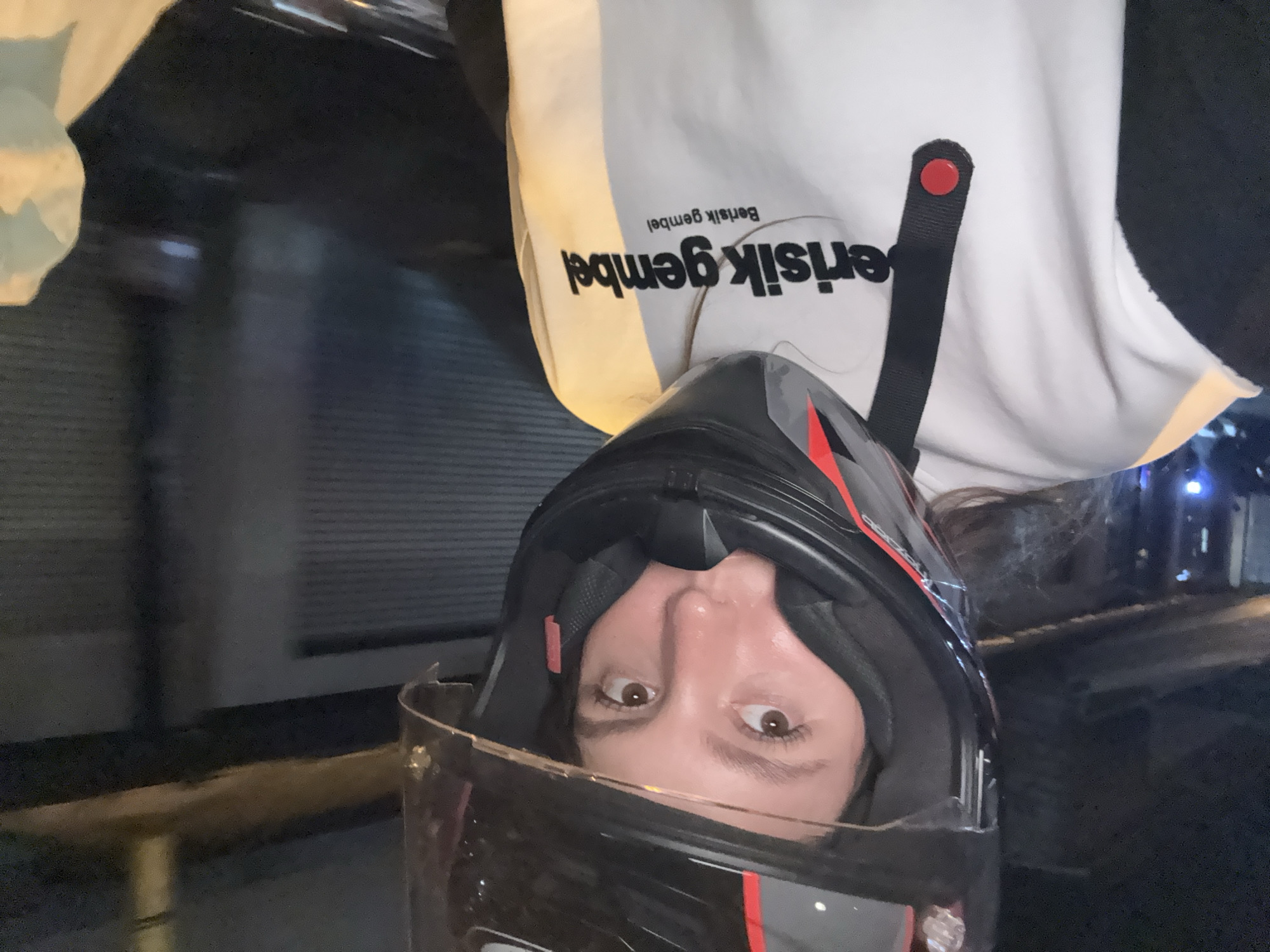 A person wearing a motorcycle helmet is visible in the foreground. The person has brown eyes and is looking directly at the camera. The helmet is black with red accents and has a transparent visor. The background appears to be an urban environment at night, with dark buildings and streetlights. In the upper part of the frame, the person is wearing a white garment with the text "Berisik gembel". The picture may have been taken in Ubud District, Indonesia.