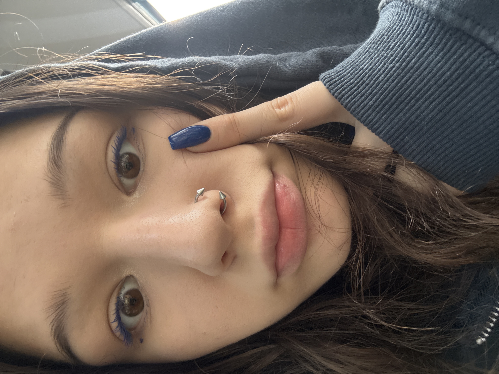 The image shows a close-up, selfie-style portrait of a young woman wearing a dark hooded sweatshirt. She has brown hair and is wearing blue eyeliner and a silver septum piercing. Her right index finger, adorned with dark blue nail polish, is touching her cheek. The background is slightly out of focus, suggesting an indoor setting or a moving vehicle. There is no discernible indication of the location being Saint Petersburg, Russia, or any specific urban environment.