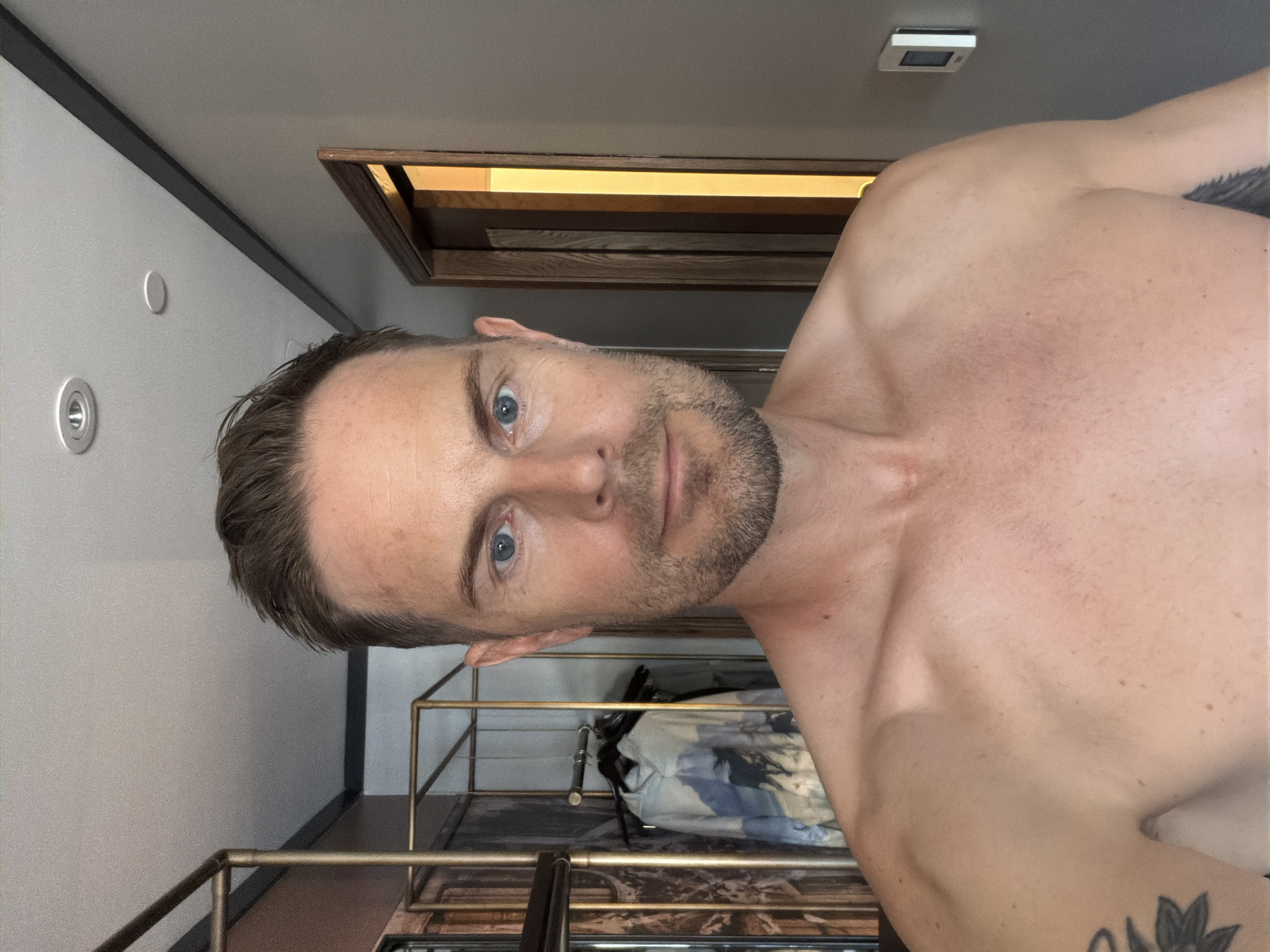 A shirtless man with short brown hair and blue eyes is looking directly at the camera. He has a light beard and a faint scar on his neck. To his left, a clothing rack with a printed shirt and other garments is visible. To his right, a doorway with warm light emanating from it is present. The environment appears to be a room with a grey wall and a ceiling with recessed lighting. There is no indication of the location in the city of Detroit, United States of America.