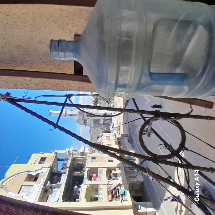 The image depicts a devastated urban landscape in Khan Yunis, Palestinian Territory, viewed from an elevated position, possibly a balcony. In the immediate foreground on the right, a large, clear, five-gallon plastic water bottle stands, partially obscuring the scene. To its left, a rusty, ornate metal railing structure is visible.
The midground features a paved street running horizontally, with scattered debris and rubble. On the left side of the street, a multi-story residential or commercial building shows severe structural damage; multiple floors are partially collapsed, exposing interiors, and laundry hangs from what appear to be damaged balconies. In the distance, a prominent mosque with a minaret is heavily damaged, its dome and portions of its walls reduced to rubble.
Several individuals are present on the street: two figures in dark clothing walk away from the viewer on the right side of the street, while another figure and a dark-colored car are visible on the left. The sky above is clear and blue, indicating daytime conditions. The overall environment conveys extensive destruction.