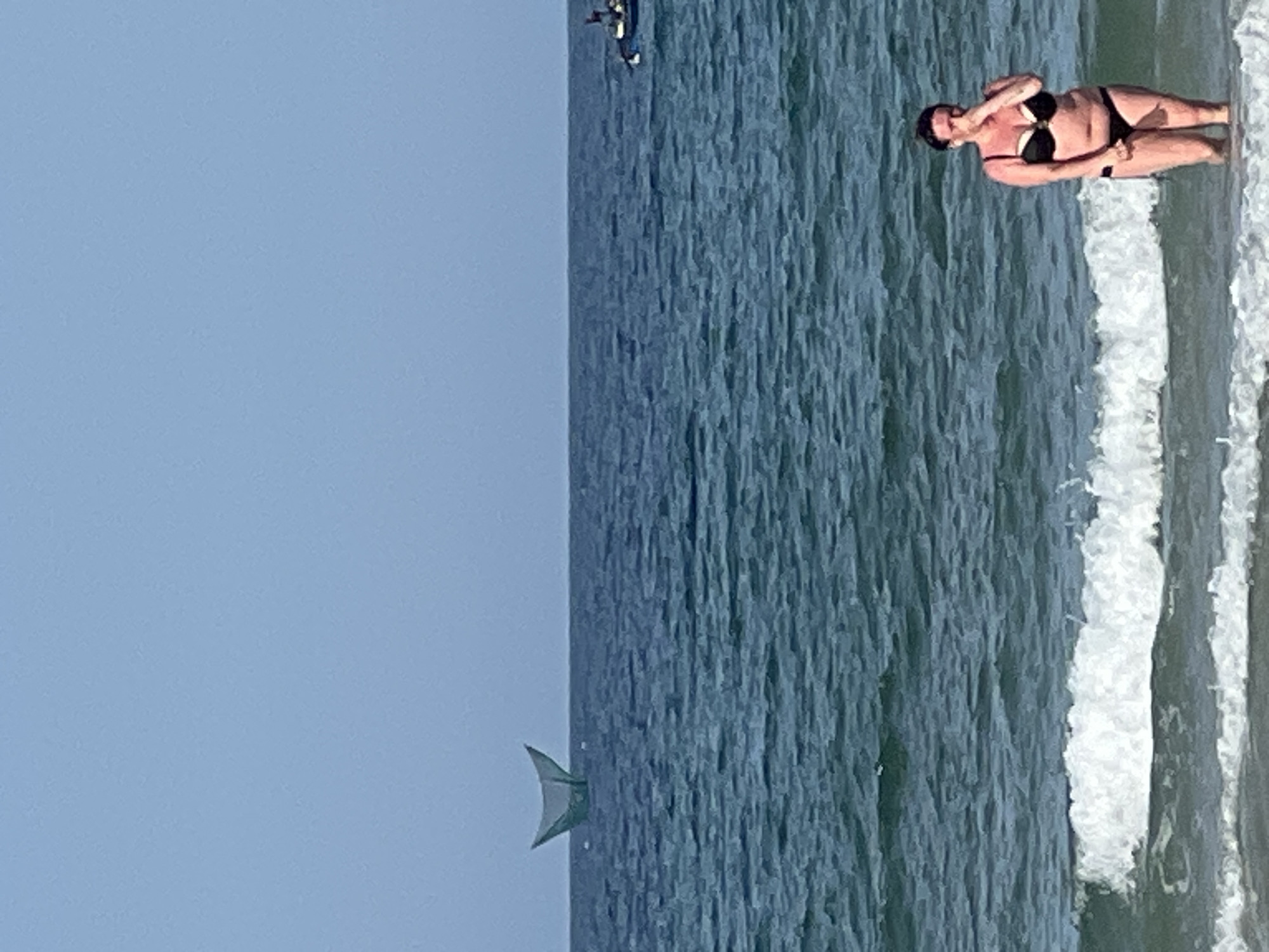 A woman stands in the ocean near the shore, wearing a black bikini. She is covering her nose and mouth with her hand. In the distance, on the left, a traditional Vietnamese fishing net is visible on the water. On the right, a small boat with people on it is also present on the ocean. The sky is clear and blue. The scene is likely on a beach in Đà Nẵng, Vietnam, given the presence of the traditional fishing net, a common sight in the region.