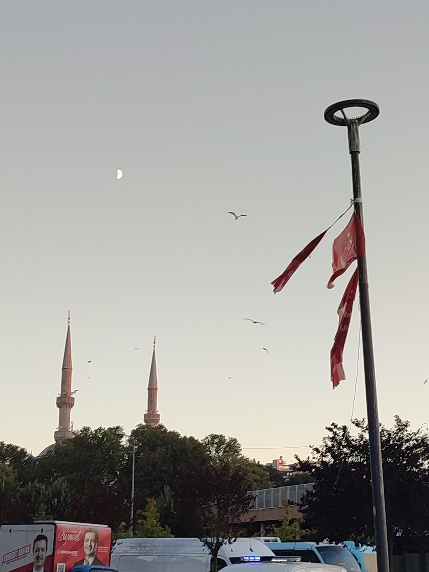 The image, taken in Üsküdar, Turkey, features a clear sky with a crescent moon visible in the upper left quadrant. In the background, the minarets of a mosque rise above a dense line of trees. In the foreground, a lamppost stands tall, adorned with three tattered red flags with white crescents and stars, likely Turkish flags. Several birds are scattered across the sky, in flight. The bottom of the image shows the top of a white van, and a portion of a billboard with images of two men and text, suggesting a political or public service announcement. The overall scene is an outdoor urban environment with a mix of cultural landmarks and modern elements.
