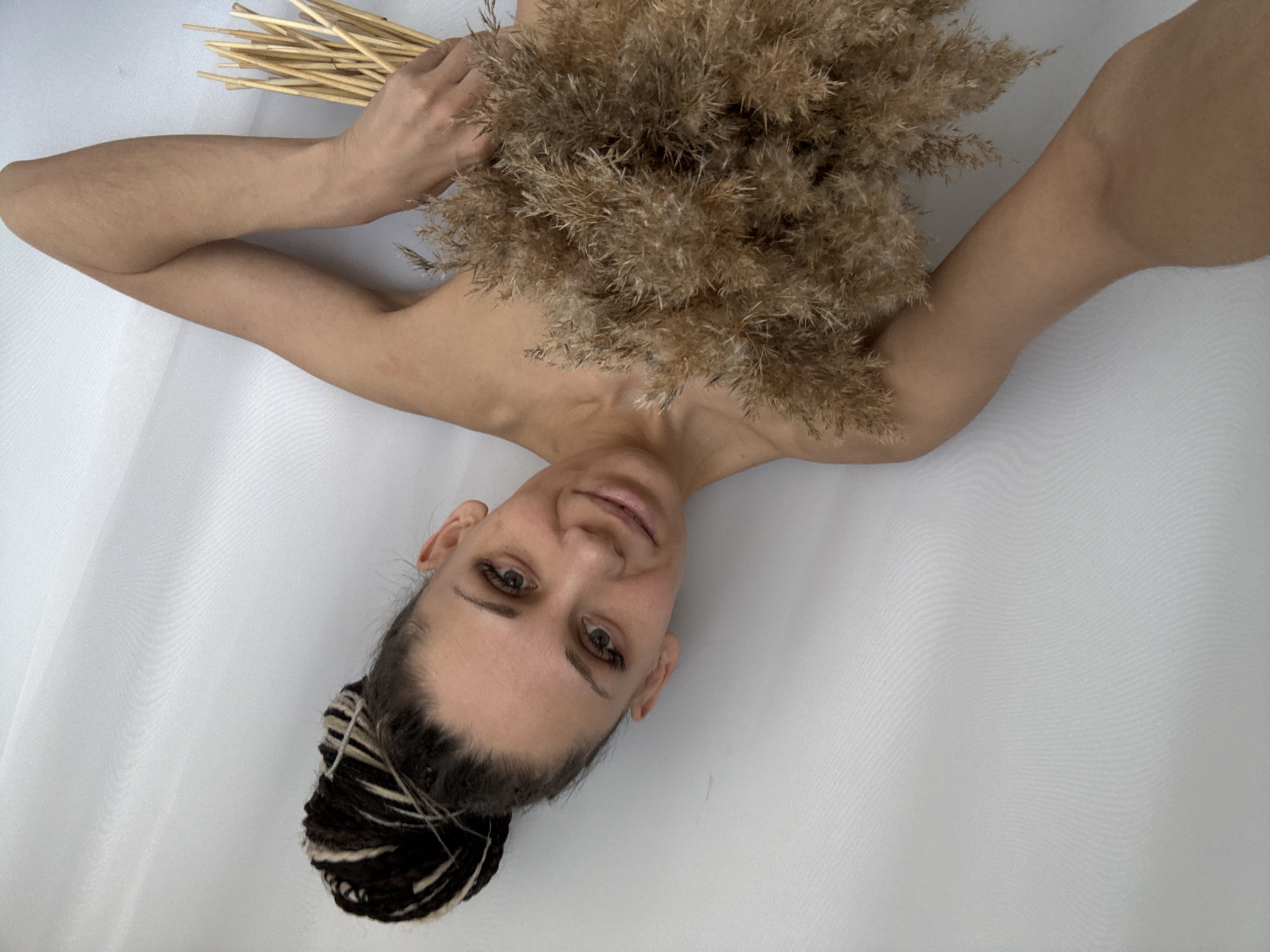 A woman lies supine on a white sheet, holding dried pampas grass and bundled straw. She has her arms outstretched. Her hair is styled in dreadlocks. The image orientation is rotated, with the woman's head positioned in the lower left corner and her feet towards the top. The background is a simple white sheet. There is no information available to confirm the location in Rusanovo, Russia, or any other specific location.