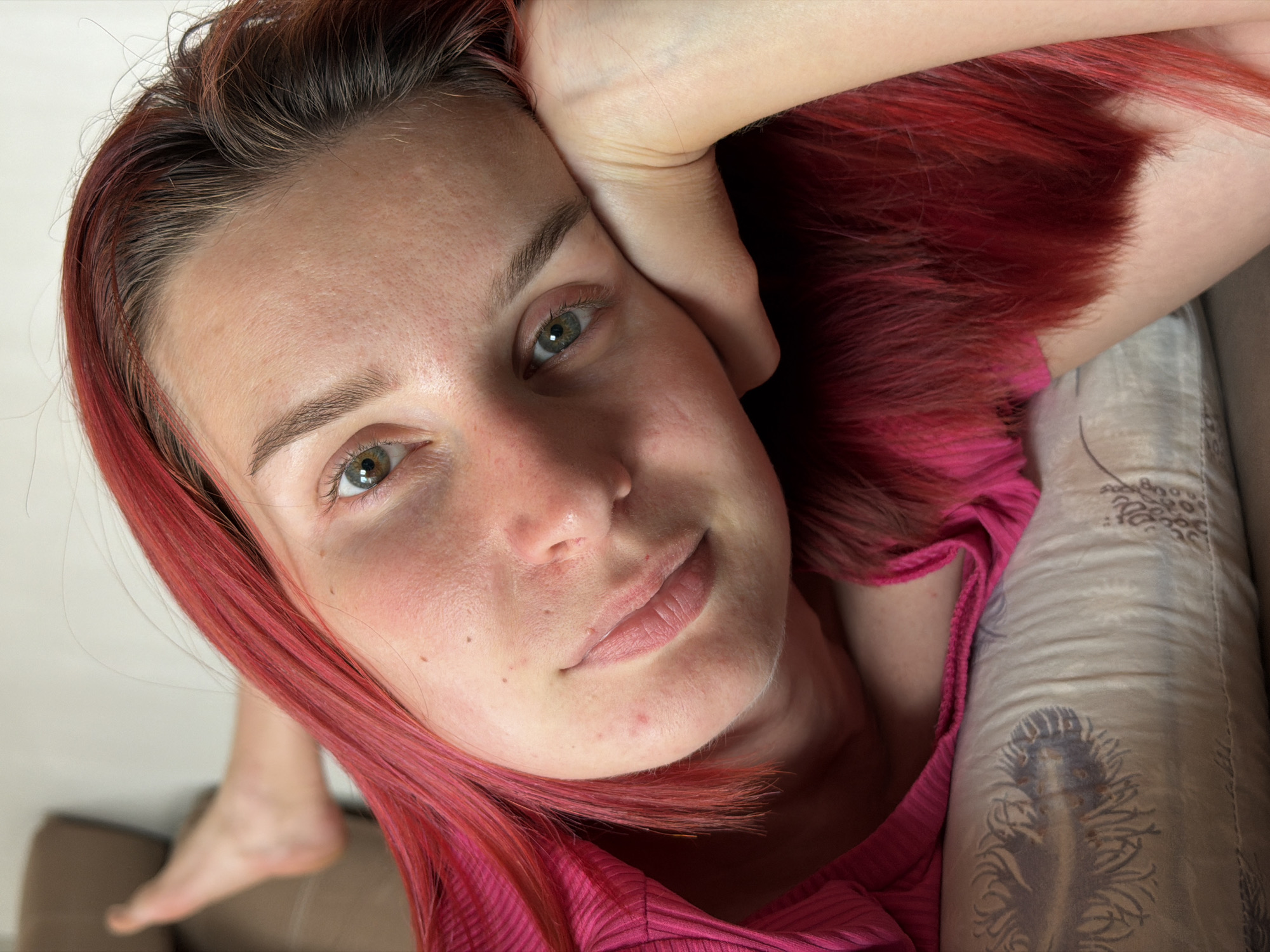 A close-up, high-angle shot depicts a light-skinned person with red hair lying on what appears to be a sofa. The person's head is resting on their hand, and they are gazing directly at the camera with a slight smile. The person is wearing a pink ribbed shirt. Part of their foot is visible in the lower left corner of the frame. The patterned pillow is located to the right of the person. The location is potentially a domestic setting, but there is no clear confirmation.