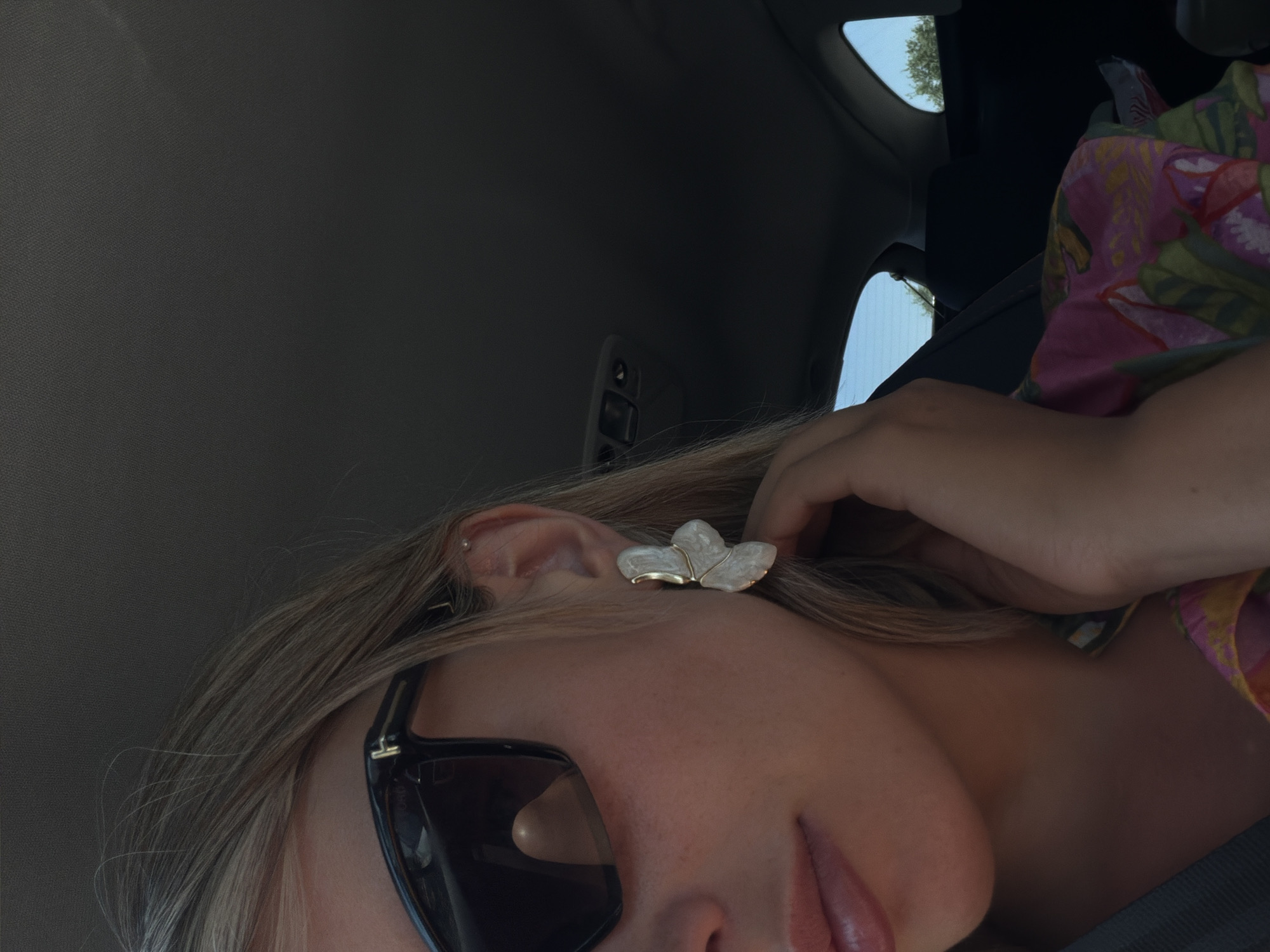 The image shows a close-up, low-angle shot of a person inside a vehicle, likely a car, given the interior ceiling and window visible. The person has blonde hair, is wearing dark sunglasses, and has on a bright, floral-patterned top. A notable accessory is a large, white, petal-shaped earring. The person's right hand is touching their hair near their ear. The background is the interior of a car, with the headliner and a window showing trees and sky outside, suggesting it is daytime and likely pleasant weather. The overall mood appears casual and relaxed, possibly indicating travel. There is no visible text in the image.