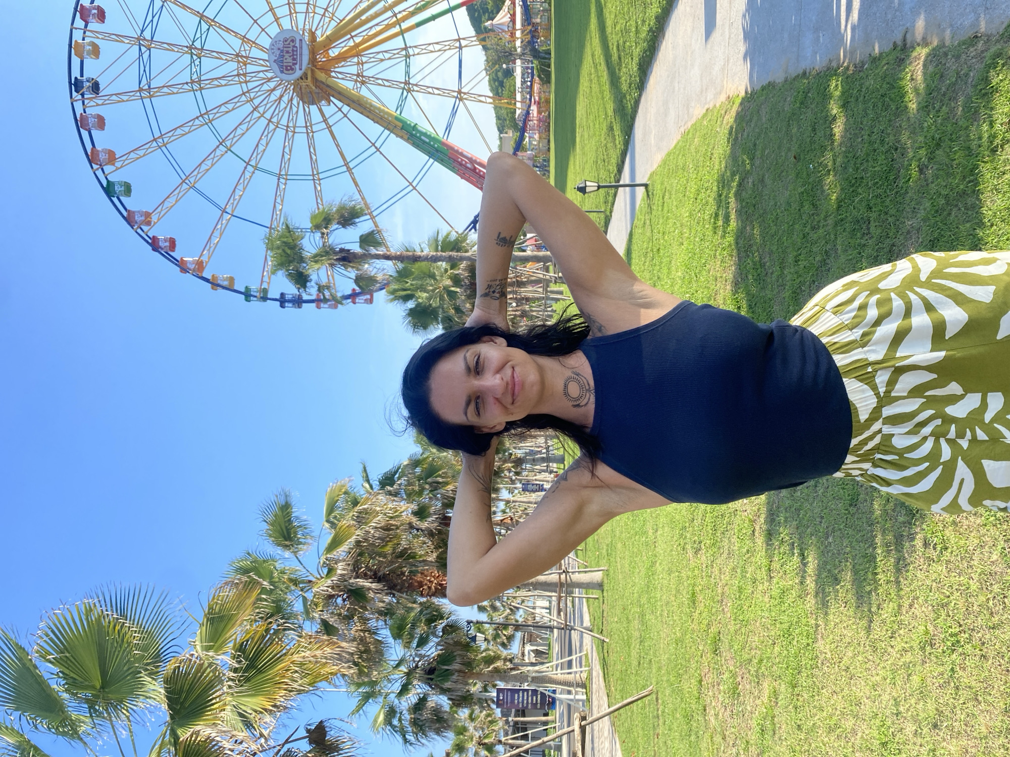 A woman stands in front of a Ferris wheel in a park in Tien Thanh Ward, Vietnam. The woman is in the foreground, facing the camera with her hands behind her head. She is wearing a dark blue tank top and a green and white patterned skirt. Behind her, a large yellow Ferris wheel with colorful gondolas is visible against a clear blue sky. Palm trees are scattered throughout the park, and a paved walkway curves through the grassy area. In the background, there are some buildings and other amusement park attractions. The overall scene depicts a sunny day at an outdoor entertainment venue.