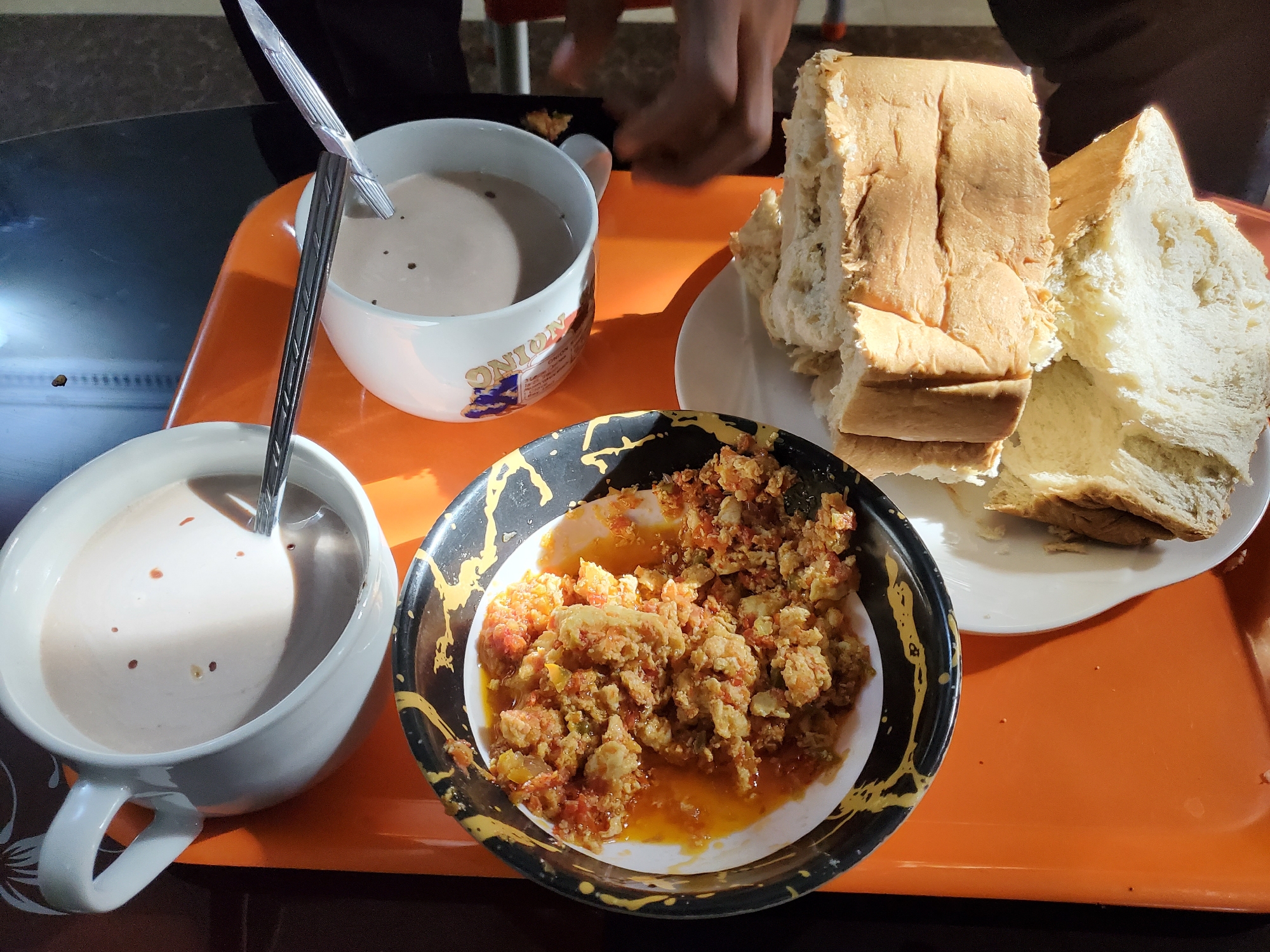 The image displays a meal laid out on a bright orange tray, likely for breakfast or a light lunch. The setting appears to be indoors, possibly a home or a casual eatery in Obosi, Nigeria, as suggested by the strong, directional natural light casting shadows on the table, indicating morning or early afternoon.
Two white mugs, each containing a light brown beverage, possibly tea or cocoa, are present. One mug clearly shows a metal spoon resting inside, while another spoon is partially visible in the second mug. The beverage has small, dark particles floating on its surface. One of the mugs features a graphic design, where the word "ONION" is clearly visible.
Adjacent to the mugs, a white plate holds several slices of what appears to be freshly baked white bread. The bread is light-colored with a soft texture. In the center of the tray, a black and white bowl with a distinctive yellow splatter pattern contains a dish of scrambled eggs. The eggs are mixed with what looks like diced tomatoes and possibly peppers, cooked in oil, creating a reddish-orange hue.
A person's hands are visible in the upper background, suggesting someone is either preparing to eat or has just placed the meal. The overall scene depicts a simple yet filling meal.
