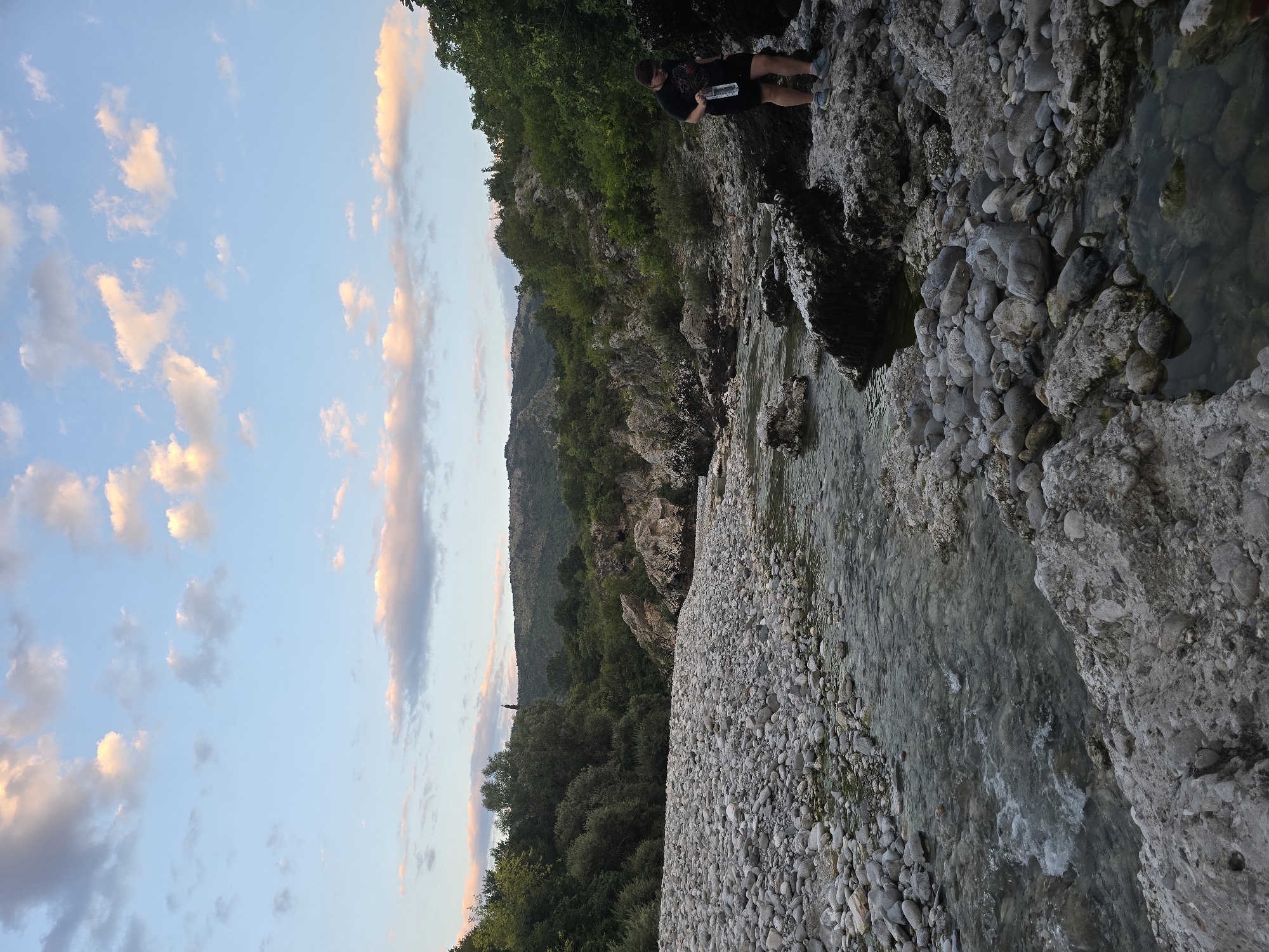 A vertical, eye-level shot captures a natural landscape with a person. The scene is set in the city of Bioče, Montenegro.
The foreground is dominated by a dry riverbed filled with smooth, grey stones and a shallow, clear stream flowing through it. The water reflects the sky and the surrounding vegetation. On the right side of the riverbed, a person stands on a large, dark rock. They are wearing a black t-shirt and shorts, holding a clear plastic bottle in their right hand. Their head is tilted down as if looking at something on the ground.
The middle ground features more rocky terrain interspersed with lush green trees and bushes. The vegetation extends up the slopes of rolling hills in the background. The hills are covered in dense, dark green foliage, creating a textured backdrop.
The sky above is a soft blue, with scattered clouds tinged with pink and orange hues, suggesting it might be early morning or late evening. The lighting is soft and diffused, casting subtle shadows. The overall atmosphere is serene and natural.