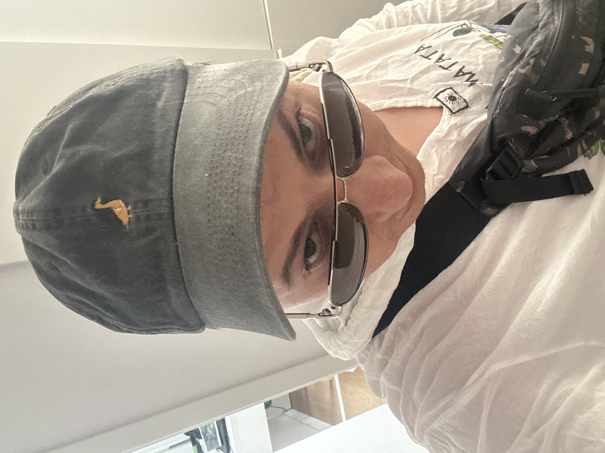 A person wearing a grey baseball cap with a small orange duck logo, sunglasses, and a white wrinkled shirt is looking down at the camera. They are also wearing a black bag with a camouflage pattern across their chest. The background appears to be an indoor setting with white walls and a doorframe visible on the left side. The image does not provide any specific information that would allow it to be placed in Benimàmet-beniferri, Spain.