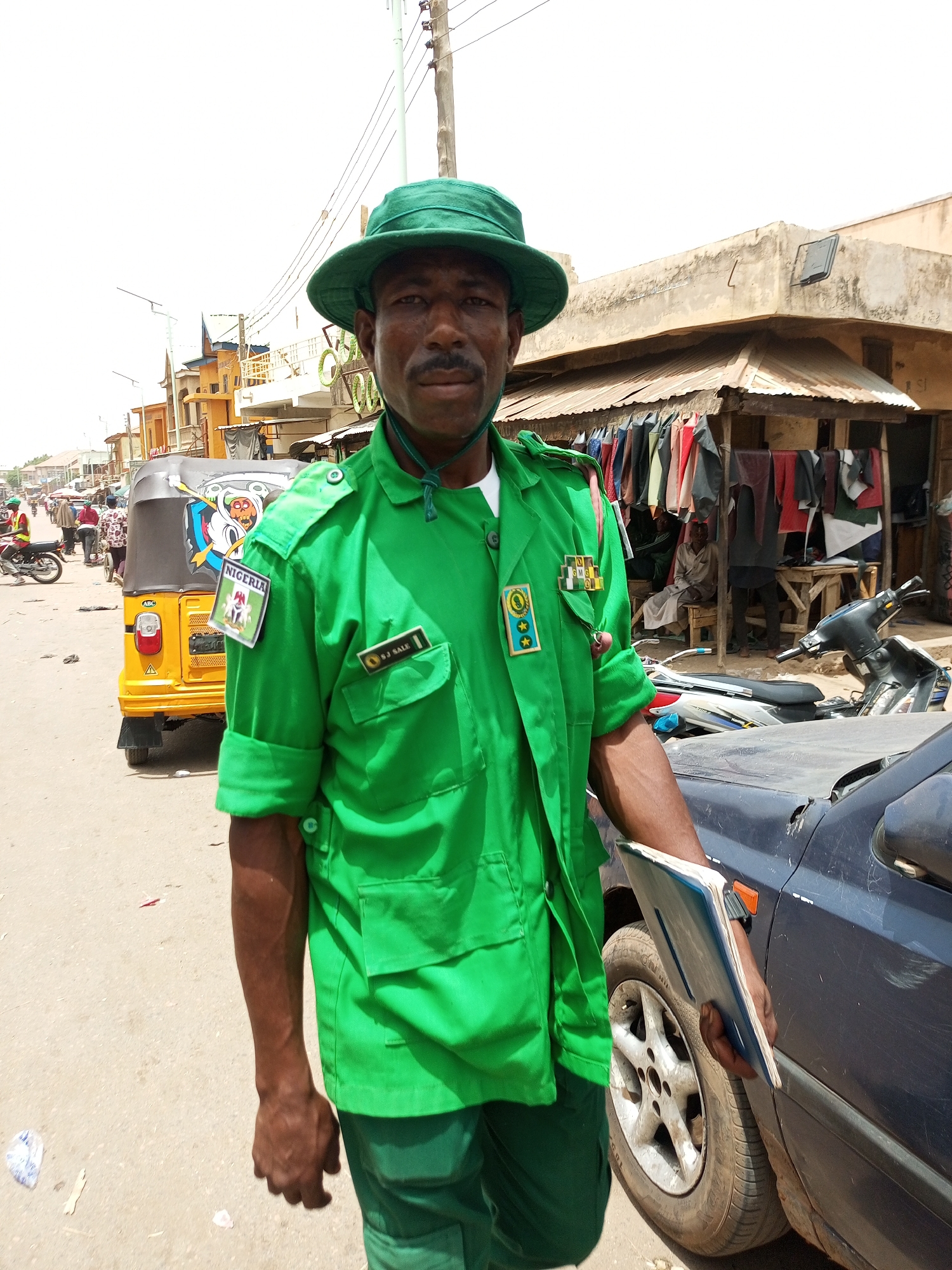 Arawa, Nigeria: A man in a green uniform and matching hat stands on a paved street, holding a blue notebook. The uniform has patches indicating "Nigeria" and a name tag, "B.J. SALE." Behind him, a yellow auto-rickshaw with colorful artwork is parked. A dark-colored car is partially visible to his right. In the background, people are walking on the street, and a building with clothing hanging outside is visible. A motorbike is parked near the building. Overhead power lines stretch across the scene.