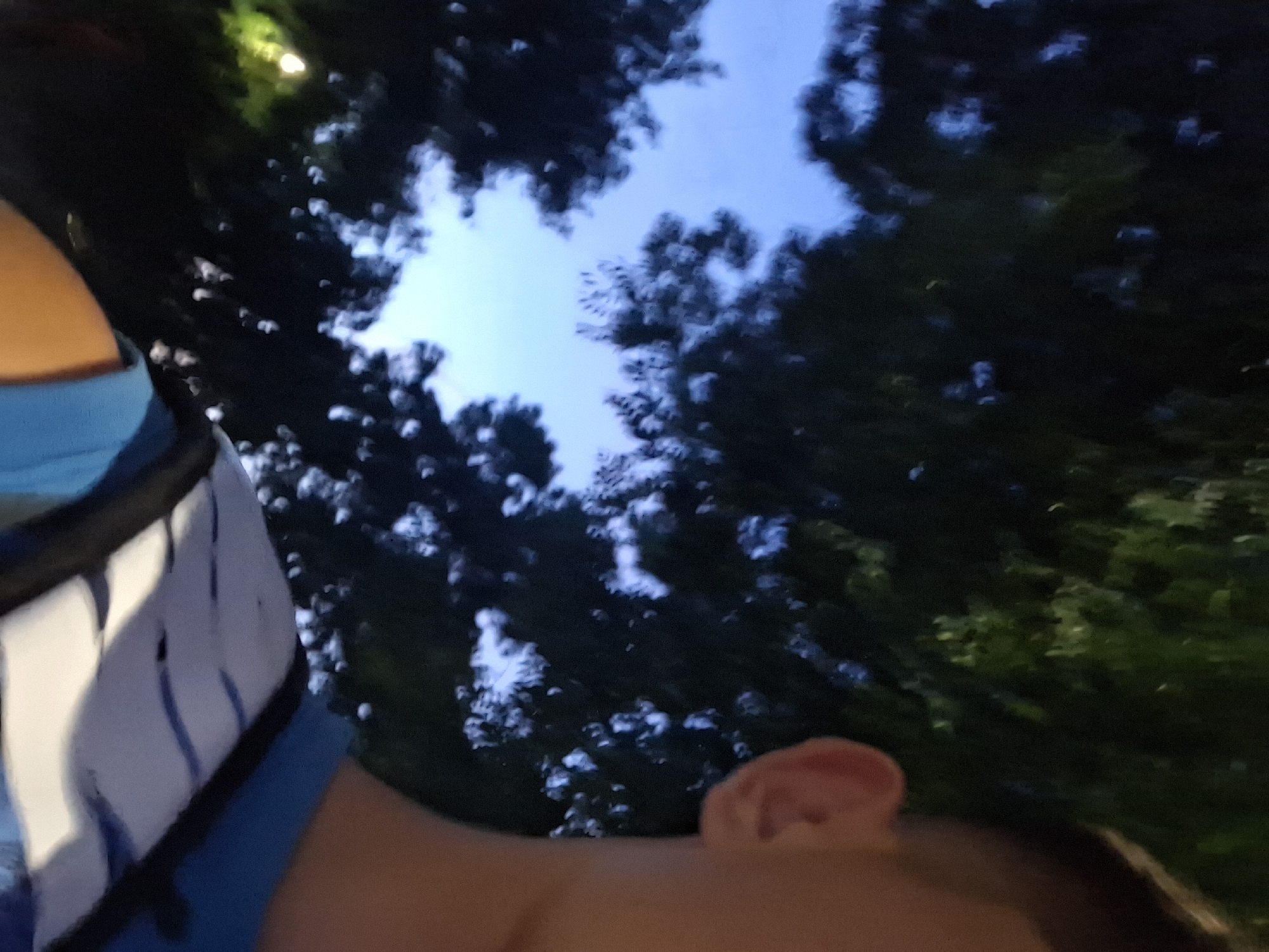 The image shows a close-up, partially blurred view of a person's head and shoulder from behind, with a blue shirt and a backpack visible. The background consists of dark green foliage and a pale blue sky, suggesting an outdoor setting during twilight. A bright, out-of-focus light source is visible in the upper left corner. The person's ear is clearly visible on the right side of the frame. There is no discernible information about the location in the city of Varna, Bulgaria.
