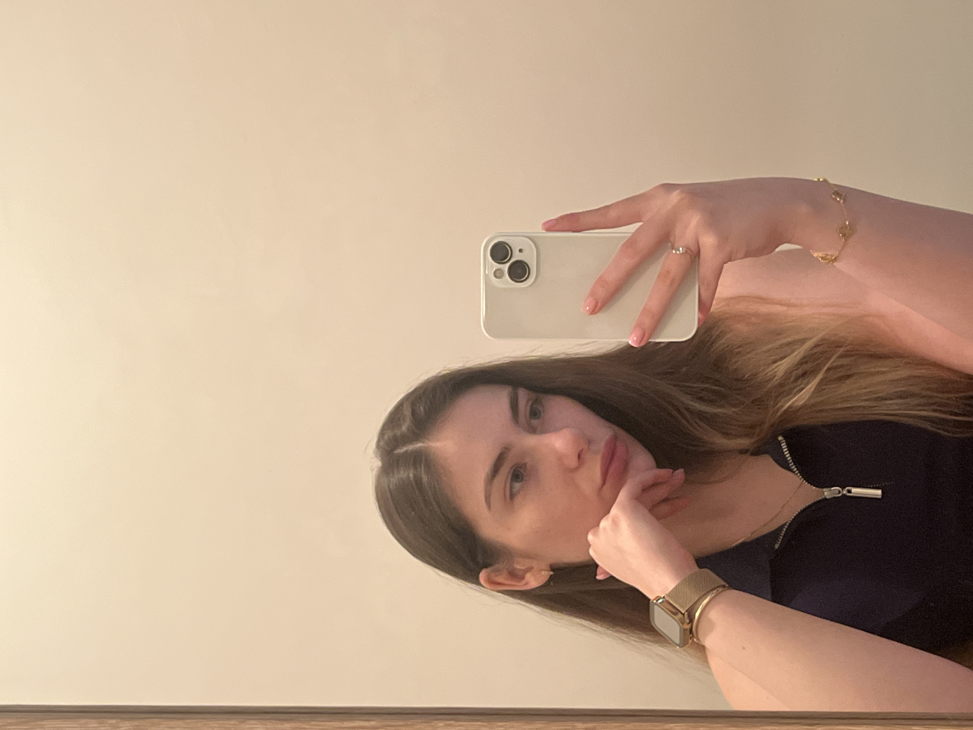 A young woman is captured in a mirror selfie, with the image rotated approximately 90 degrees counter-clockwise. She has long, dark brown hair, parted in the middle, and a direct gaze towards the camera with a neutral to slightly pensive expression. She wears a dark top, possibly a quarter-zip, with a silver-toned zipper visible at the neckline.
In her right hand, she holds a light-colored smartphone (likely white or light grey, possibly an iPhone due to the dual camera lenses) to take the picture. Her right hand also features a delicate gold-toned bracelet with small charms on her wrist and a silver-toned ring on her ring finger. Her left hand is positioned near her chin, with fingers gently touching her lips. On her left wrist, she wears a gold-toned smartwatch with a mesh strap. Both of her hands display light-colored nail polish.
The background is a plain, light beige or off-white wall, indicating an indoor setting. A narrow strip of brown, possibly wooden, flooring or furniture is visible at the bottom edge of the frame. The lighting is soft and even. There is no visible text in the image.