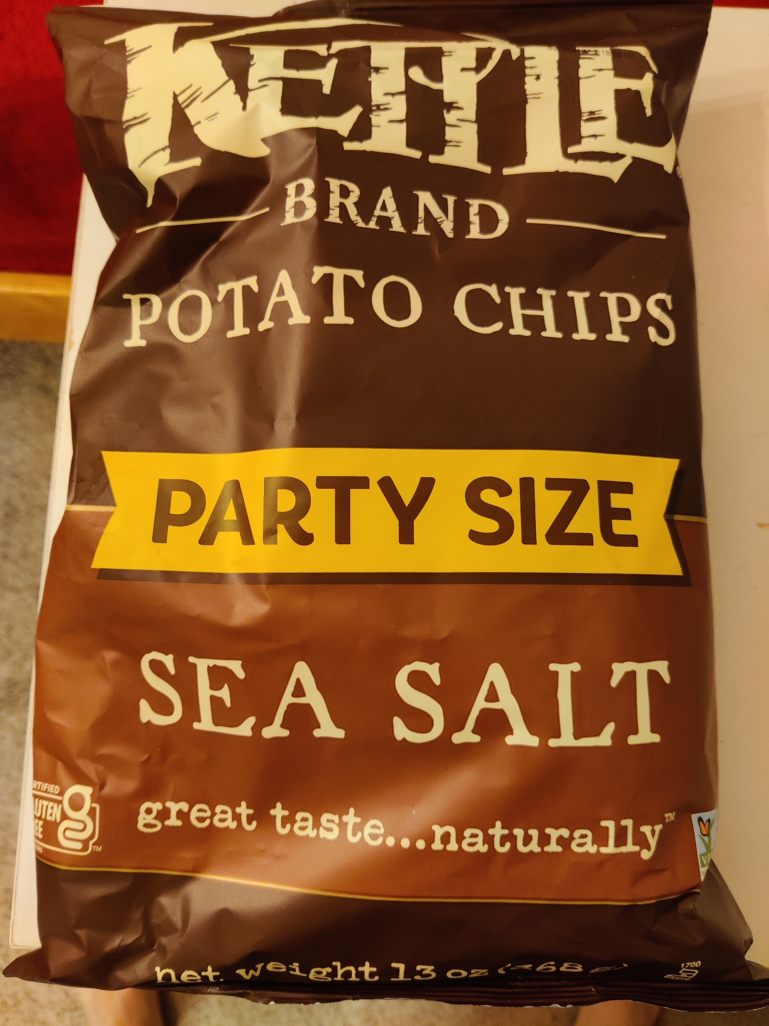 A top-down, close-up shot shows a bag of Kettle Brand Sea Salt Potato Chips. The bag is predominantly brown with off-white lettering. The brand name "KETTLE" is at the top, followed by "BRAND POTATO CHIPS" below it. A yellow banner across the middle reads "PARTY SIZE". Below that, "SEA SALT" is prominently displayed. A gluten-free certification logo and the text "great taste...naturally" are visible towards the bottom. The bottom edge of the bag has text indicating "net weight 13 oz (468g)" and a recycling symbol with the number 1700. The image is taken from a high angle, with the bag laid flat. The scene appears to be indoors, with a subtle red background element visible to the left. No specific location in Lower Paxton Township, United States of America, can be identified from this image.