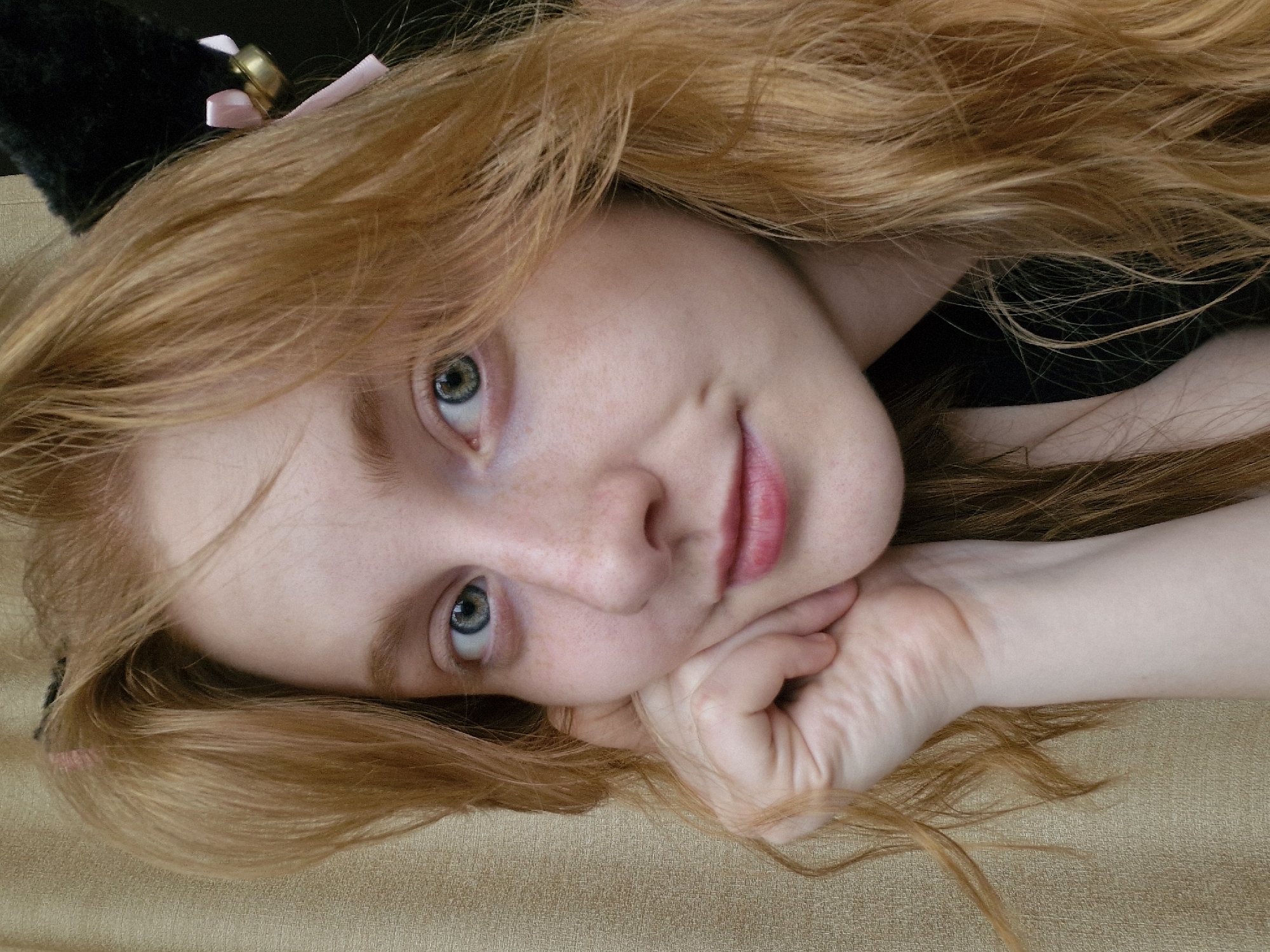 A close-up portrait features a young woman with long, wavy, reddish-blonde hair, lying down and looking towards the camera. Her head is tilted to the right, resting on her right hand. She has fair skin, light freckles visible across her nose and cheeks, and striking blue-green eyes. Her lips are slightly parted in a subtle, natural smile.
She is wearing a pair of black, furry cat ears, with a small pink ribbon and a golden bell attached to the ear visible on the left side of the frame. A dark garment, possibly black, is visible around her upper body and neck area. Her hair spreads out around her head and across the textured, light brown or beige surface she is lying on, which appears to be upholstery or fabric. The lighting is soft and even, suggesting an indoor setting without direct harsh light. No text is visible in the image. The scene is set in Yekaterinburg, Russia.