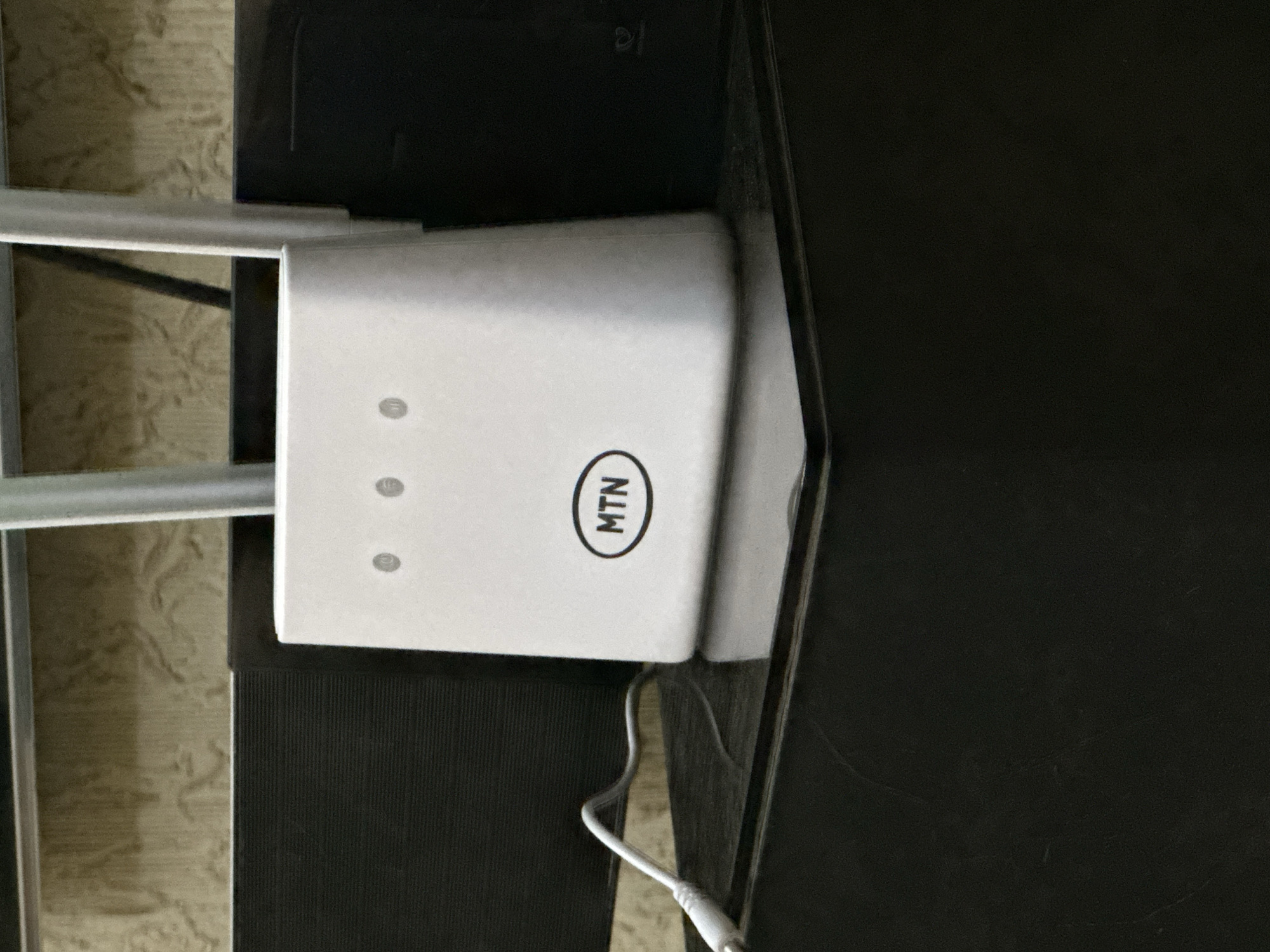 The image shows a white MTN Wi-Fi router placed on a surface. The router has three indicator lights and the MTN logo. A power cable is connected to the router. The surfaces surrounding the router are black and beige. The location is Unknown, Unknown.