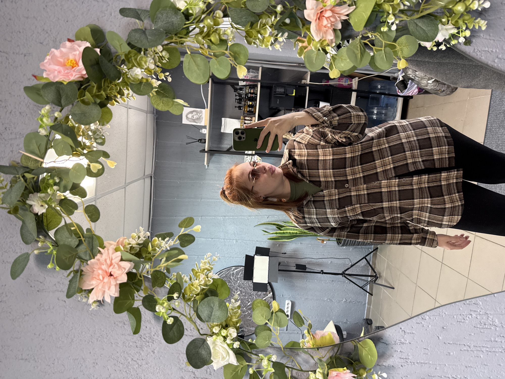 The image shows a reflection in a mirror decorated with flowers and greenery. A woman with red hair and sunglasses is visible in the mirror, holding a green smartphone, presumably taking the photograph. She is wearing a brown plaid shirt and black pants. The reflection shows a room with gray walls and a tile floor. There is a light stand, a plant, and shelves with various bottles and equipment visible in the background. The location of the image is Chaikovsky, Russia.