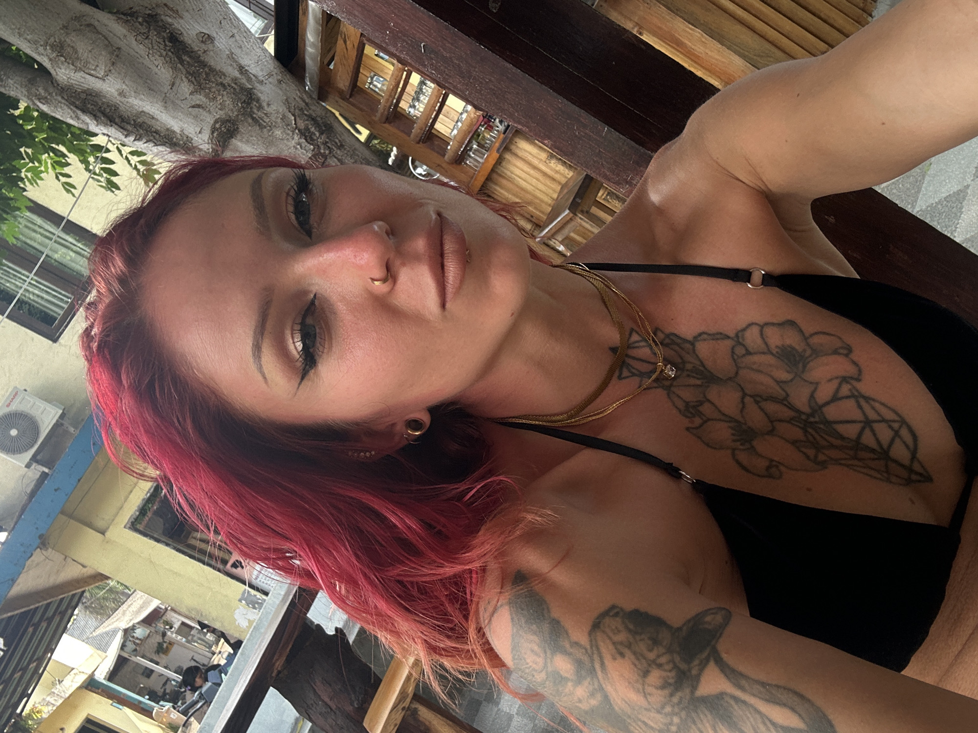 A woman with red hair and tattoos is in Unknown, Indonesia. She is wearing a black top and gold necklaces. There is an air conditioner visible, suggesting an indoor or partially covered space. She appears to be in a restaurant or similar establishment, based on the tables and background details.