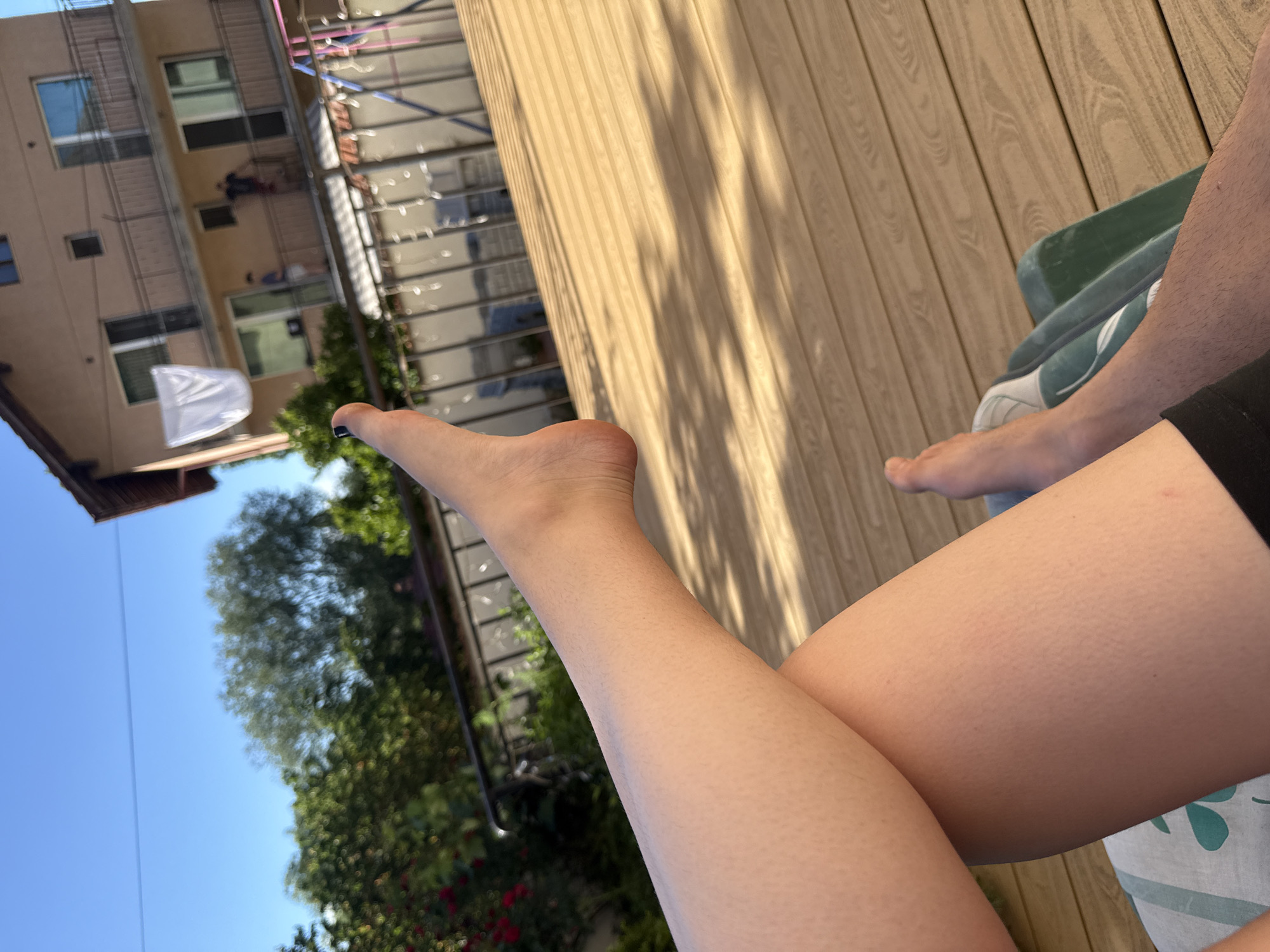 The image shows a close-up of a person's legs and feet, resting on a wooden deck. The person is lying down, with their legs crossed. The right leg is extended, and the foot is visible, wearing a white sneaker. The left leg is bent, with the thigh in focus. The skin on the legs appears to have goosebumps.
In the background, there is a three-story building with balconies, a fence, and lush green trees. A white sheet is hanging from a clothesline on one of the balconies. Two figures are visible on the balconies of the building, one standing and the other appearing to be leaning over the railing. The scene suggests a residential area, possibly a backyard or a patio, with a sunny sky overhead. The setting appears to be outdoors on a warm day.