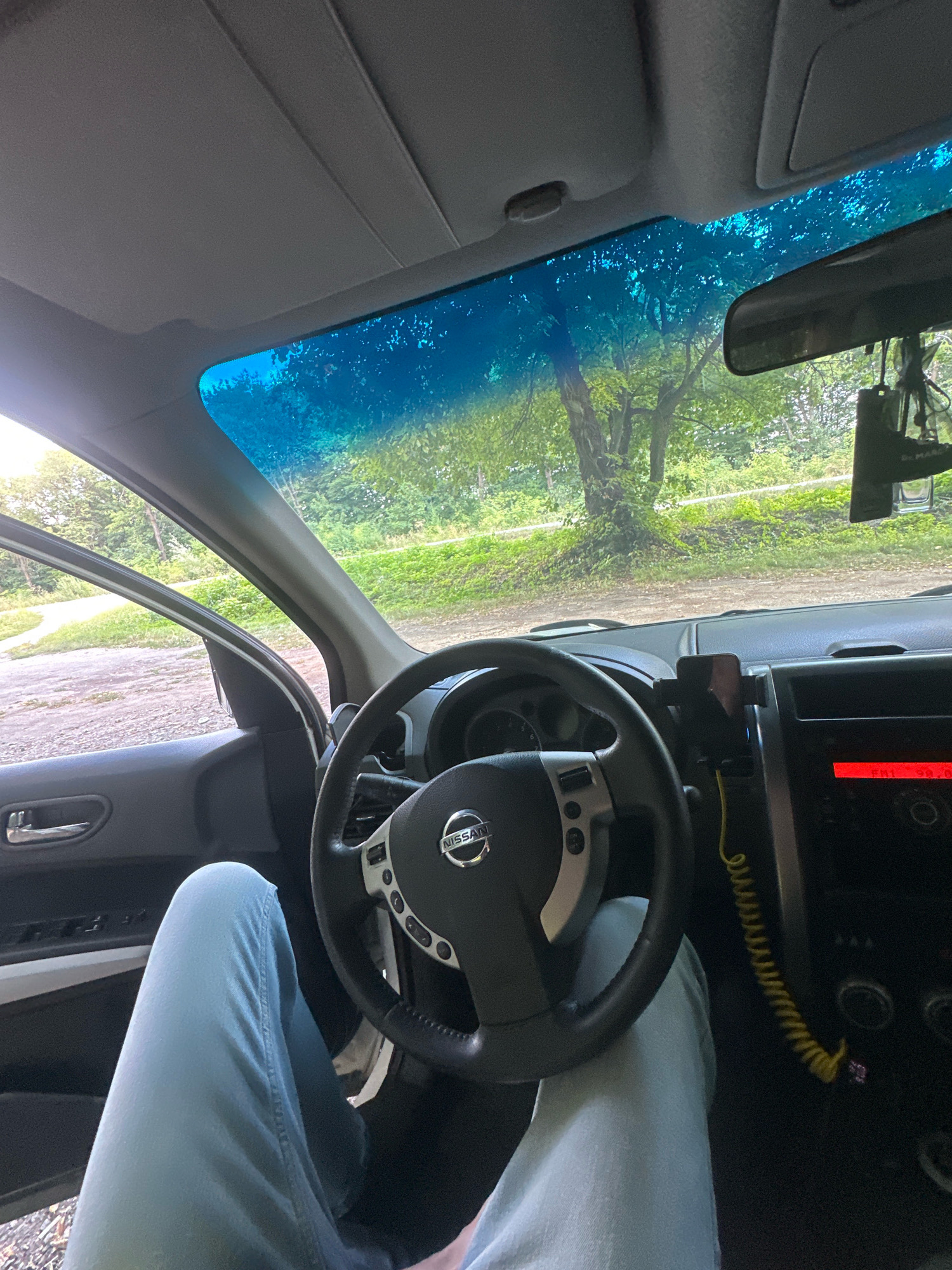 The image shows the interior of a car, looking forward through the windshield. The driver's legs, clad in light blue jeans, are visible in the foreground. A steering wheel with a Nissan logo is centered in the frame. To the right of the steering wheel, a car stereo display shows "FNL 90.20". A yellow coiled cord hangs from a phone mount attached to the dashboard. Outside the car, through the windshield and open driver's side door, a tree-lined, somewhat rural landscape is visible, with a dirt path or road in the distance. The sky above is a bright blue with some foliage reflected on the windshield. The image appears to have been taken in Shatava, Ukraine, given the context provided.