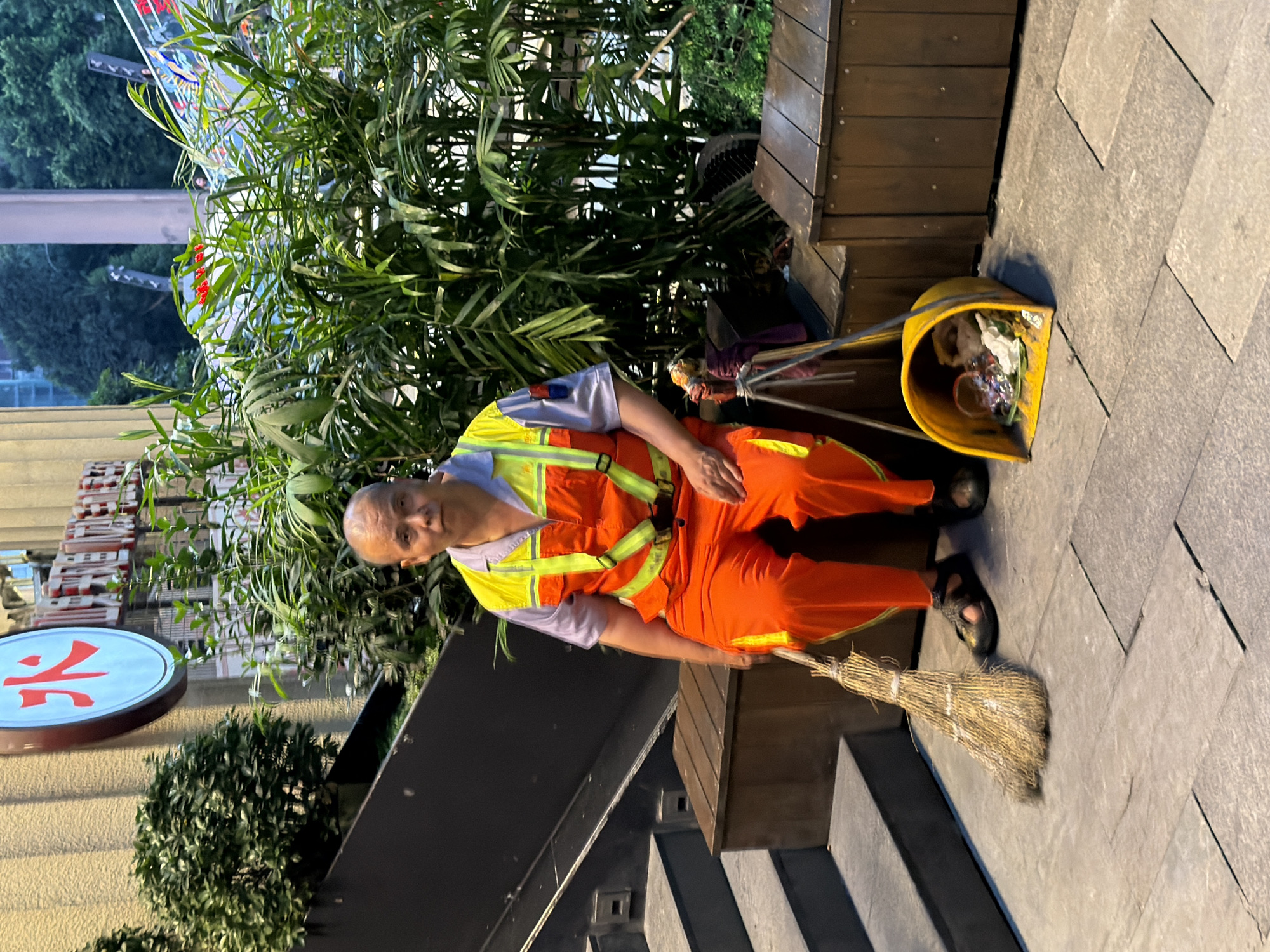 In Yuzhong District, China, a bald man wearing an orange and yellow high-visibility vest and orange pants sits on a wooden bench next to a set of stairs. He has a broom in his left hand and a yellow dustpan containing debris to his right. Lush green plants are visible behind him, with a building and trees in the background. The ground is paved with large, light-colored stones.