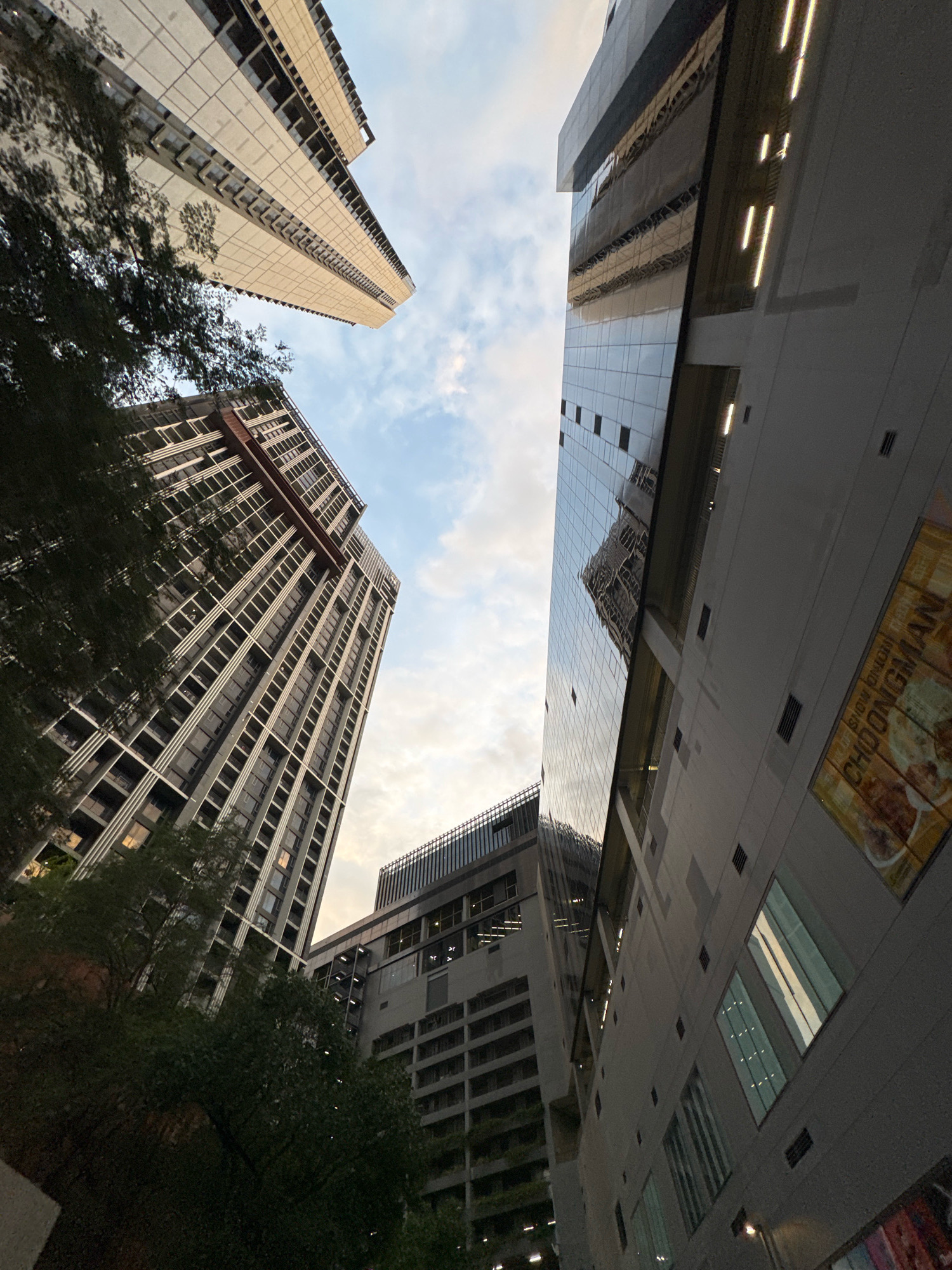 The image depicts a low-angle view of modern skyscrapers in Bangkok, Thailand. The perspective is looking upwards, showcasing the architectural styles of several tall buildings. On the left, a residential or office building with a facade of light-colored panels and numerous windows is visible, partially obscured by dark green foliage in the foreground. To its right, another skyscraper with a grey facade and grid-like window pattern rises. Dominating the right side of the frame is a contemporary building with a sleek, reflective glass exterior that mirrors the sky and other structures. This glass facade features linear lighting elements near the top. Further in the background, another building with a darker grey facade and horizontal window bands can be seen. A sign with Thai script, partially legible as "CHONGMAN," is affixed to the exterior wall of the glass building on the right. The sky above is a mix of blue with scattered clouds, suggesting it is daytime, possibly late afternoon or early evening due to the lighting conditions. No people or specific actions are depicted in the scene.