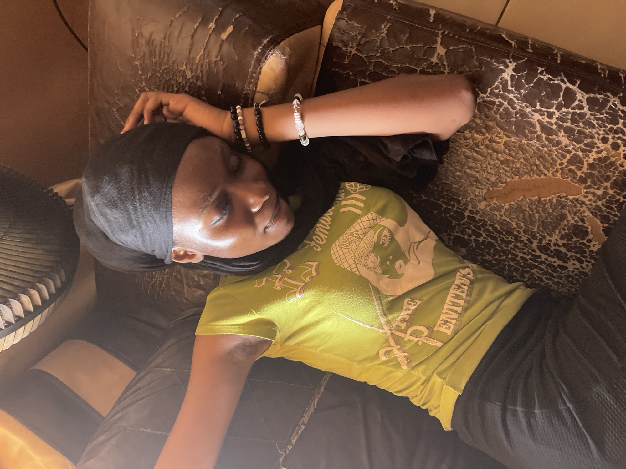 A young woman wearing a black head covering and a light green t-shirt with a graphic design is lying on an old, worn, dark brown leather couch. She is wearing bracelets on her right wrist. The couch shows signs of age and wear, with cracking and peeling leather. Part of an electric fan is visible in the lower left corner. The location, Unguwar Gabas, Nigeria, is contextual information that isn't directly discernible from the visual elements of the image alone.