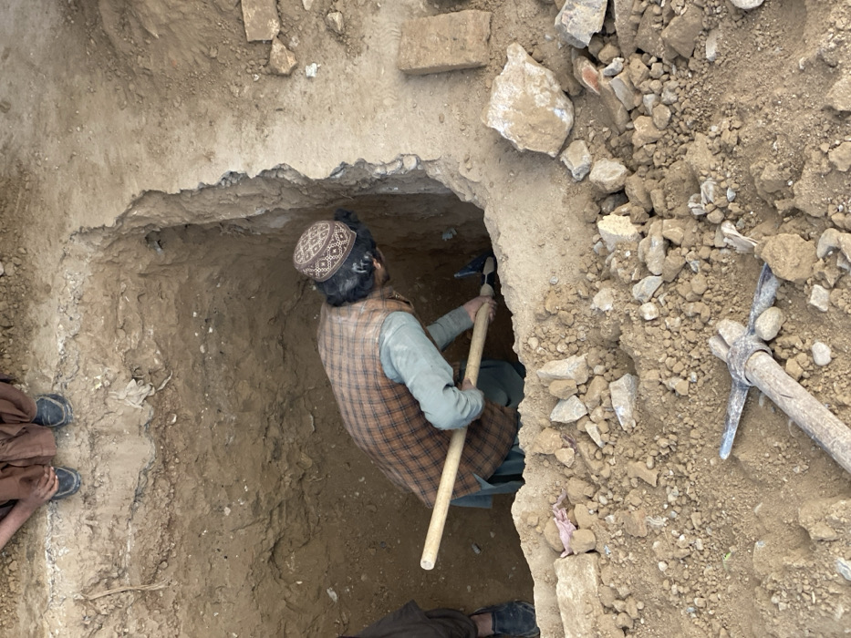 The scene depicts an active excavation within a dry, earthen environment in Kandahar, Afghanistan. A male subject is positioned inside a roughly rectangular pit, estimated to be 1.5 to 2 meters deep, actively digging with a pickaxe. He wears a light-colored tunic, a brown plaid vest, dark trousers, and a patterned cap, with his head bent forward, focused on the digging action. The lower legs and feet of a second individual, dressed in dark shoes and brown trousers, are visible standing on the undisturbed ground surface at the top edge of the pit, observing. The surrounding ground is composed of loose, rocky soil with scattered debris, including a second pickaxe lying horizontally in the immediate foreground. The excavated pit walls show variations in soil composition and some embedded detritus.