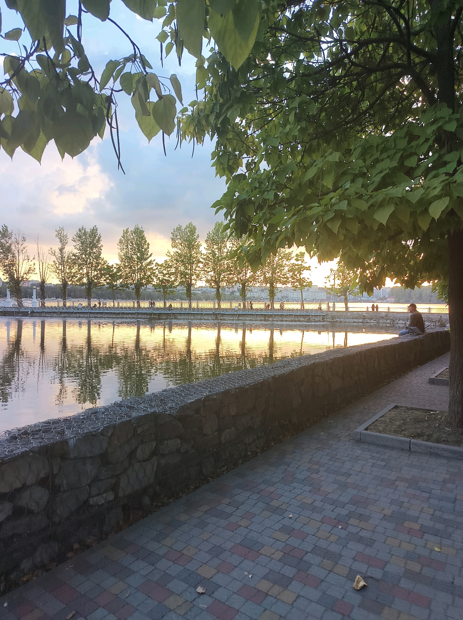 This image captures a sunset view of the Ternopil lake, specifically the embankment near the city center. In the foreground, a woman is sitting on a stone wall, facing away from the camera, looking out at the water. The wall is constructed of large stones and is reinforced with wire mesh. A paved walkway with interlocking brick patterns runs alongside the wall. Trees with lush green leaves frame the scene from the upper left and right, with some of their branches and foliage casting shadows on the ground and water.
The lake itself is calm, reflecting the warm hues of the sunset and the silhouettes of trees and buildings on the opposite bank. A row of trees lines the far embankment, their reflections clearly visible on the water's surface. In the distance, a city skyline is visible, bathed in the golden light of the setting sun. The sky above is a mixture of soft clouds and clear blue, with the sun's glow creating a warm ambiance. The overall atmosphere is serene and peaceful, a typical evening scene at Ternopil lake.