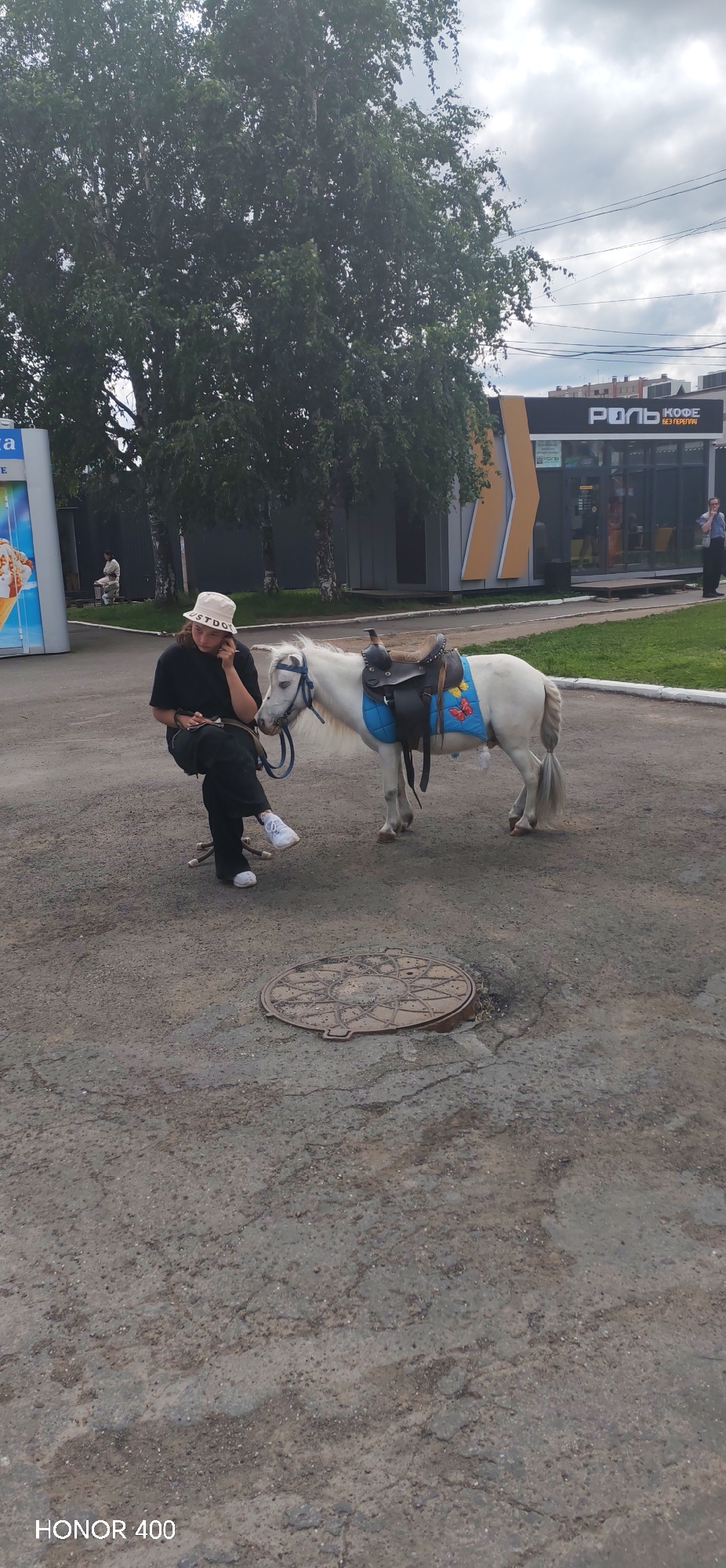 A person wearing a black t-shirt, black pants, white sneakers, and a white bucket hat is sitting on a chair outdoors, talking on a cell phone and holding a pen. Next to the person is a small white horse with a saddle and a blue blanket with butterfly decorations. The horse is tethered. The scene appears to be in a city, with a building in the background that has signage that includes the Russian words "РОЛЬ" and "КОФЕ". There are trees and power lines visible in the background, and the ground is paved. The overall environment suggests a casual, outdoor setting, possibly a public space or a street. The image is watermarked with "HONOR 400".