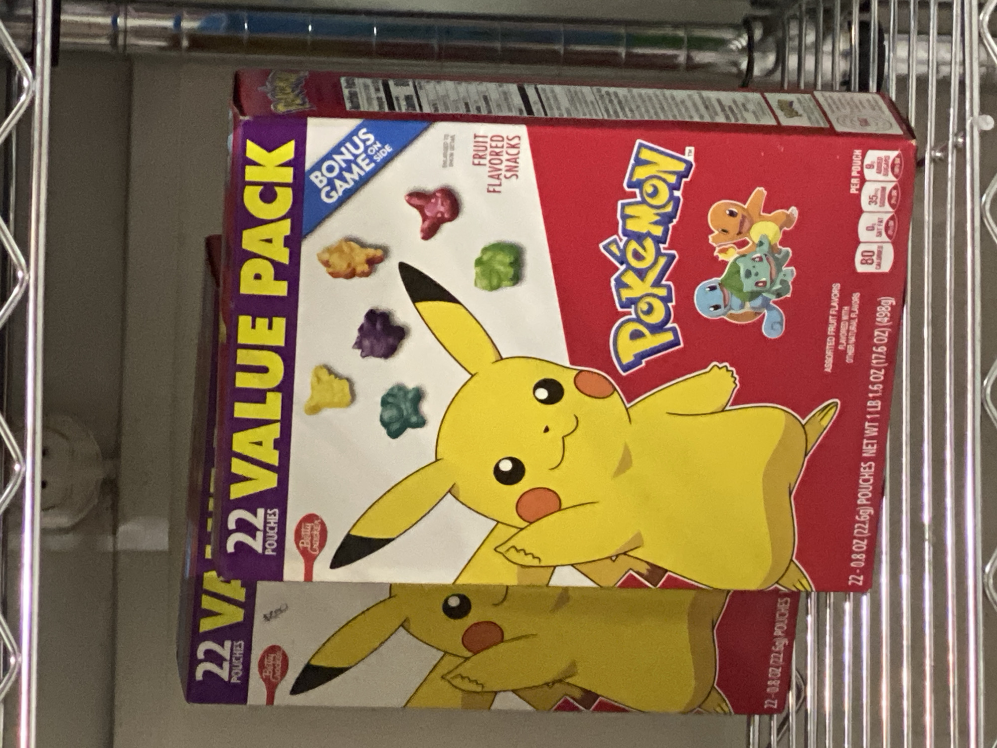 The image shows a box of Pokémon fruit snacks on a metal rack, likely a storage or shelving unit. The box is red with a large image of Pikachu on the front. The side of the box, colored purple, reads "22 Value Pack." Text on the box also indicates it contains "Fruit Flavored Snacks" and has "Assorted Fruit Flavors." The box is likely located in a pantry or storage area. The image does not contain any information on location, so city of San Bernardino Tlaxcalancingo, Mexico cannot be confirmed.