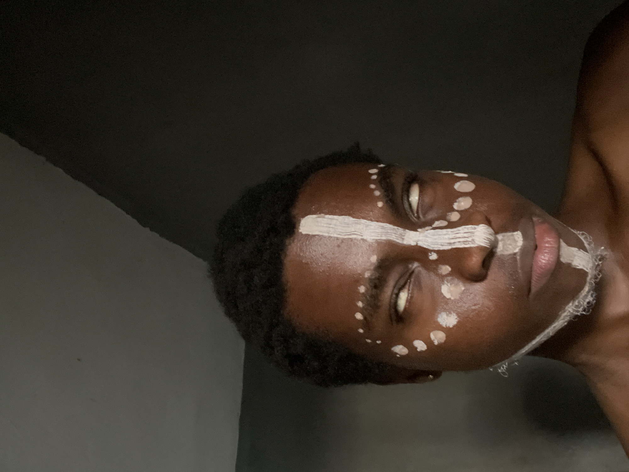 A medium shot depicts a young man with dark skin and short curly hair, positioned with his face angled downwards to the left. He is wearing white paint or similar substance in a linear pattern across his forehead and on his chin, with dots accentuating his cheeks and near his eyes. The background appears to be an interior space with dark grey walls. The image provides no evidence linking it to Onitsha, Nigeria.