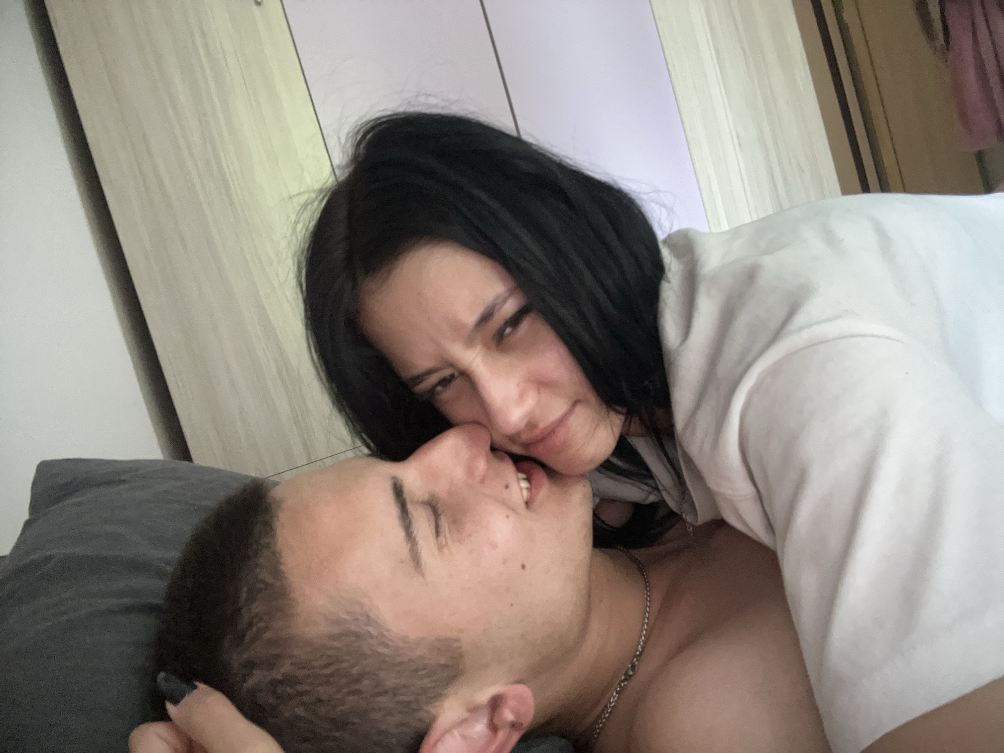 A woman with dark hair is lying on top of a man in bed. The man has short hair and is wearing a chain necklace. The woman is wearing a white shirt. The location appears to be a bedroom. There is no evidence to support the location being Burgas, Bulgaria.